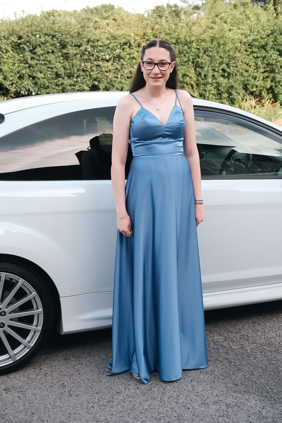 Young person in blue formal gown standing next to white car outdoors.