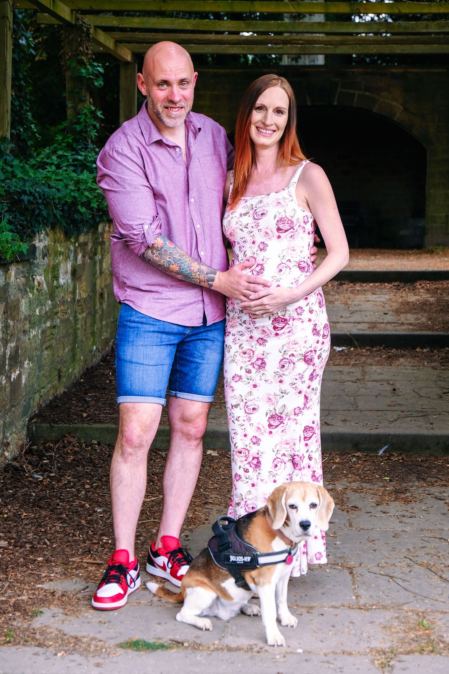 A couple wearing pink and floral attire poses with their beagle dog in an outdoor setting.