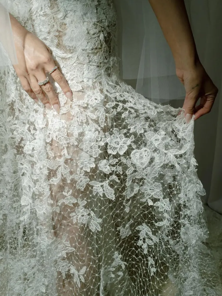 Close-up detail of intricate white lace wedding dress fabric being held to show delicate floral pattern and texture.