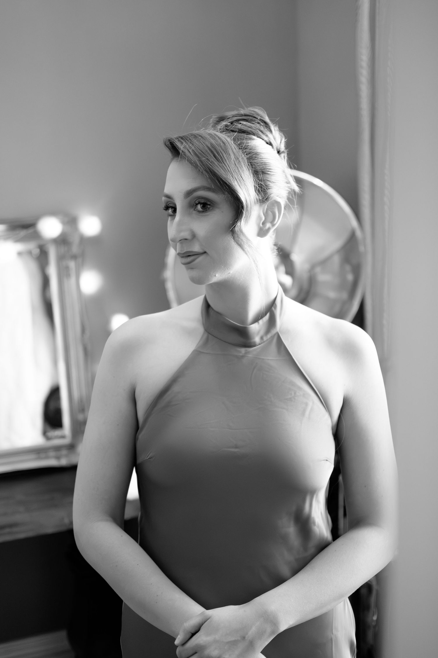 Black and white photo of an elegant woman in a halter dress with an updo hairstyle, posing in front of a lit vanity mirror  at dunston hall