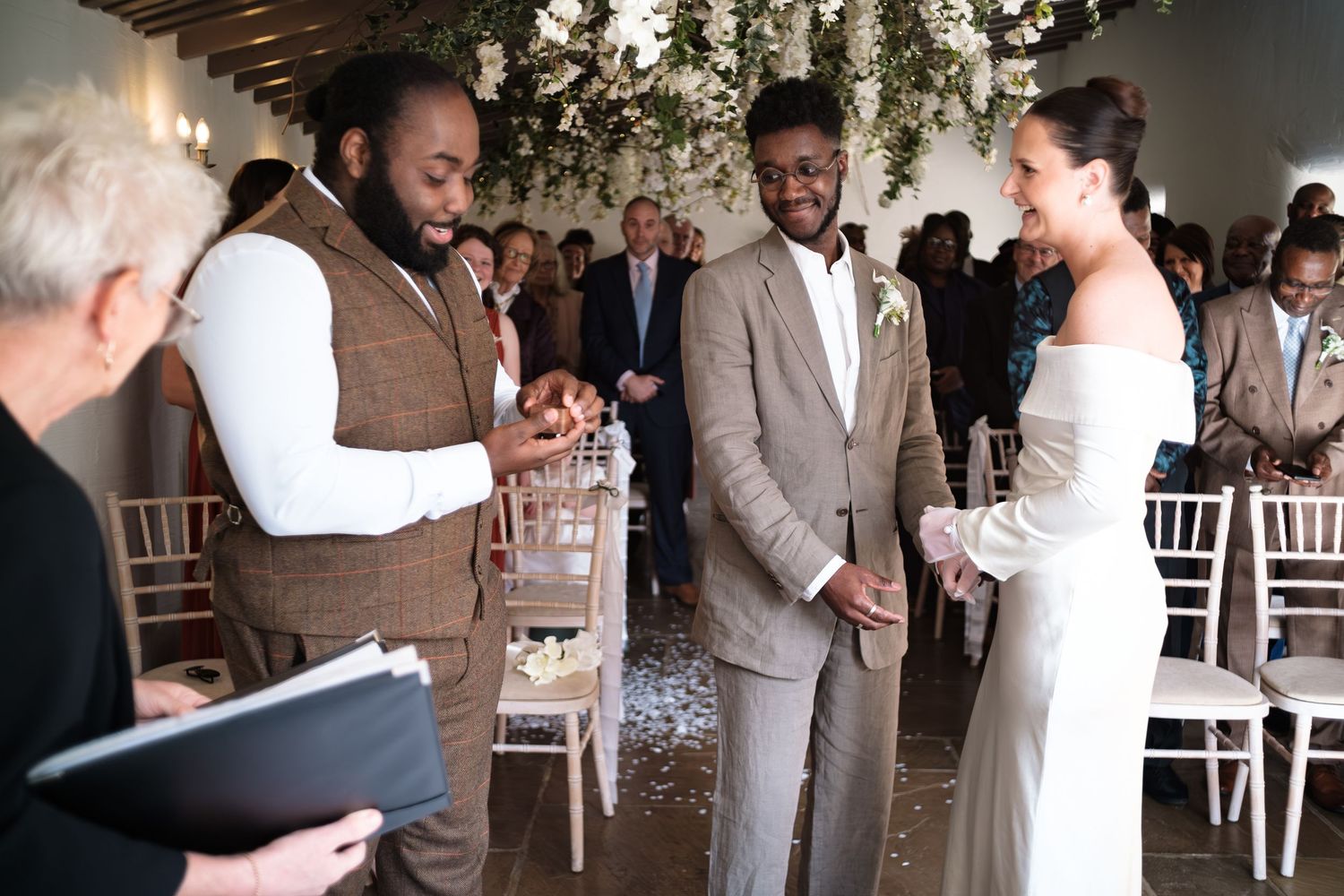 Wedding ceremony with groom in brown tweed suit, best man, and bride in white off-shoulder gown at elegant floral venue.