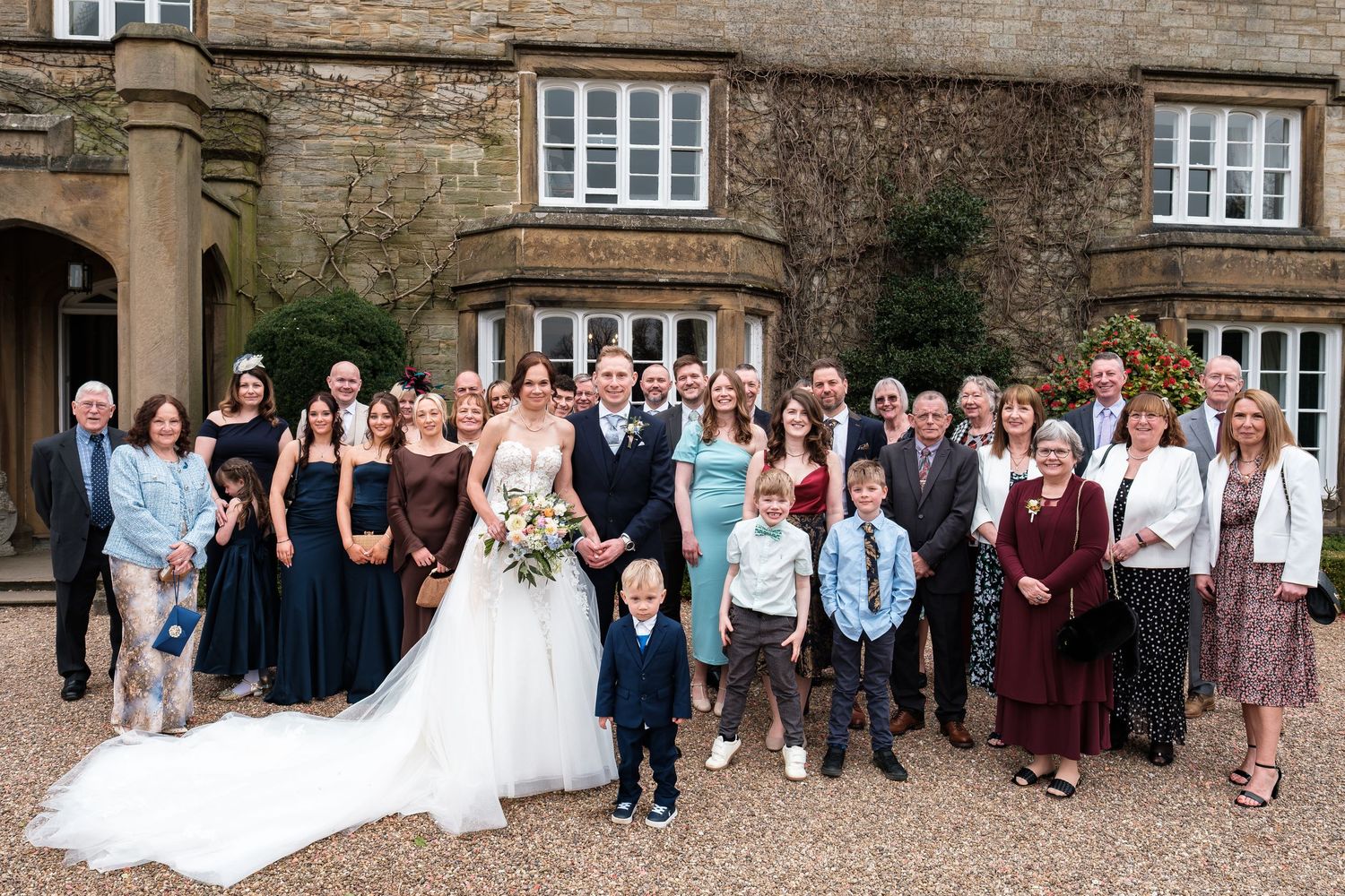 Wedding party group photo with bride and groom surrounded by family and friends outside a historic stone manor house.