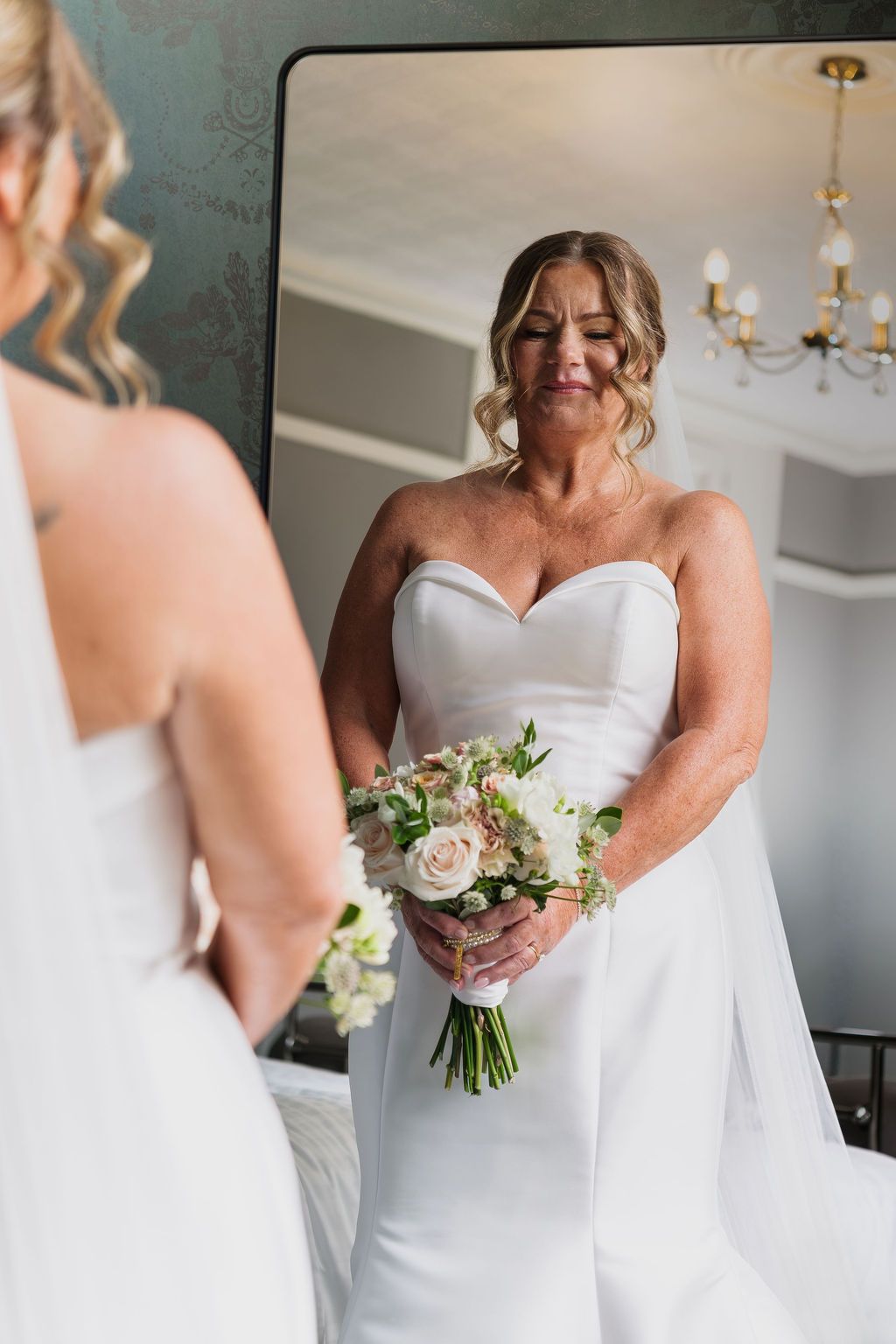 Bride in white strapless gown holds blush bouquet, gazing at her reflection in a large mirror before her wedding.