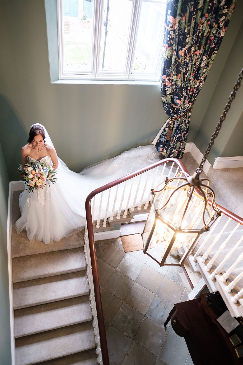 Bride descending the staircase seen from above, bouquet trailing, iron lantern in frame