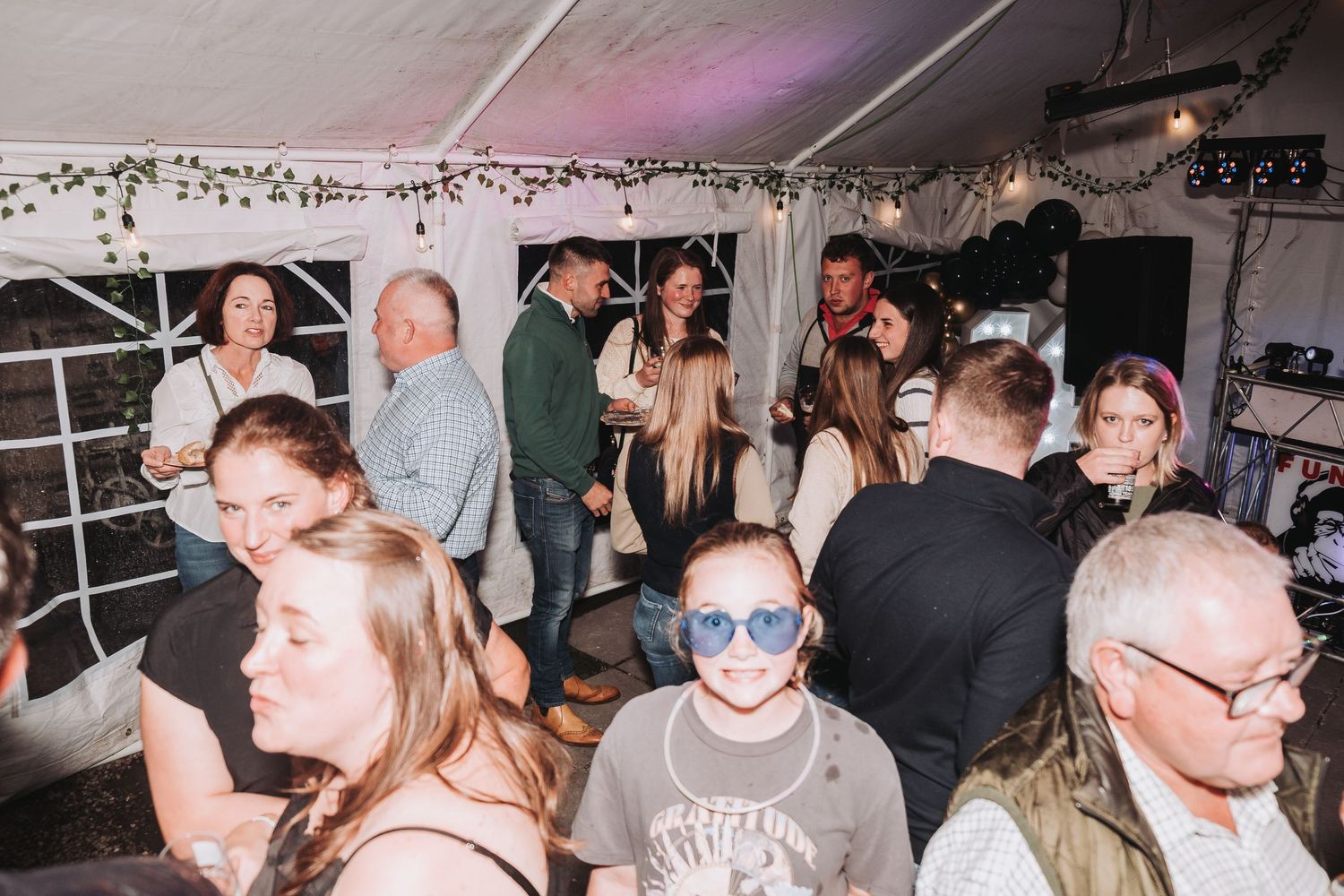 A crowded party scene under a white tent with people dancing and socializing.