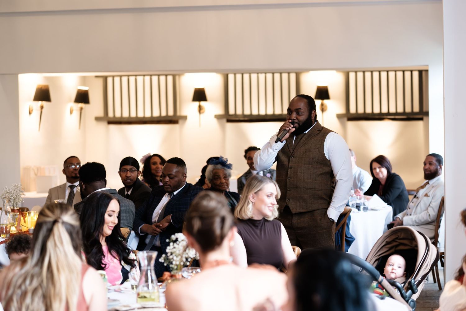 A man in a vest speaks to guests at an elegant wedding reception dinner in a well-lit dunston hall estate
