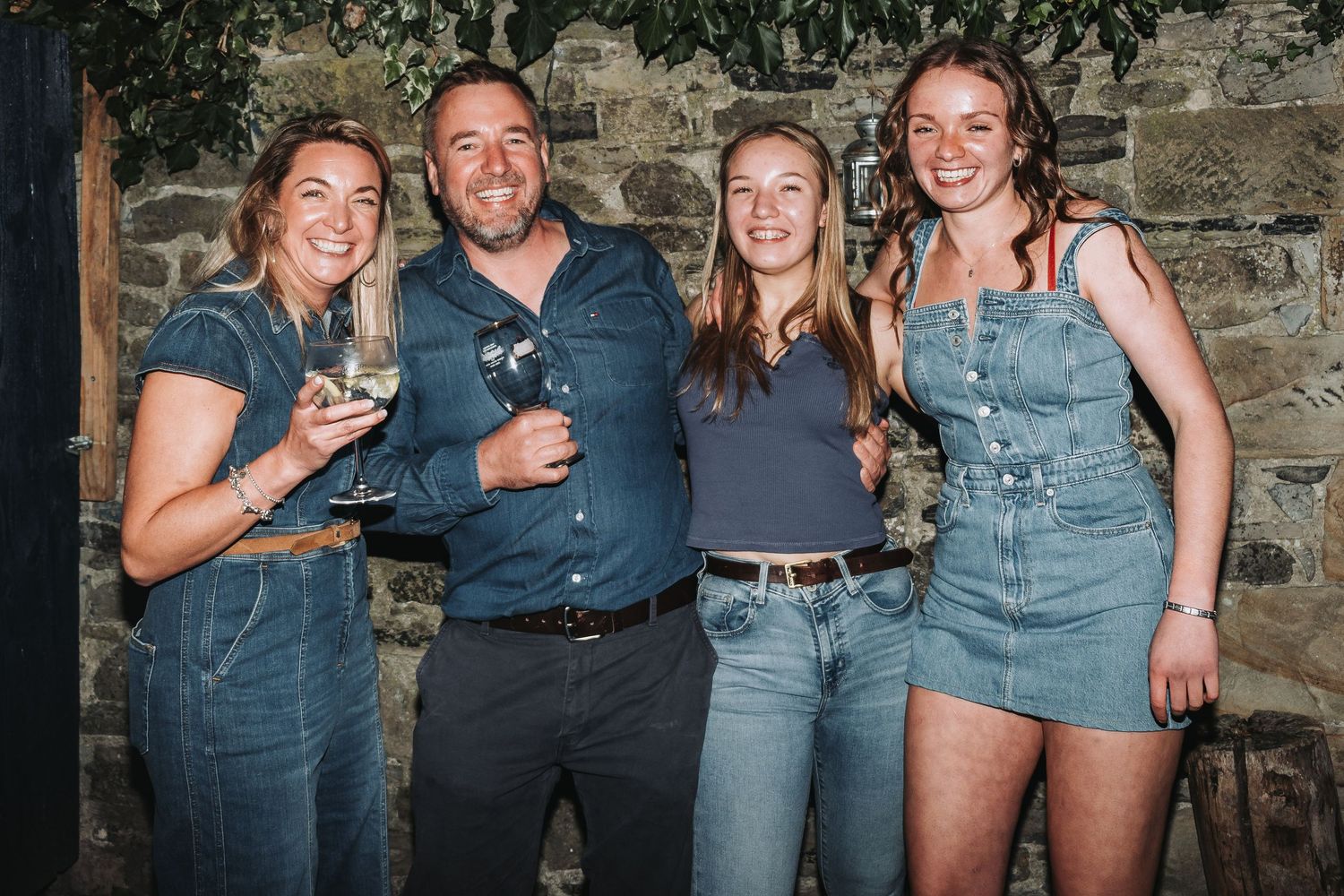 A group of friends in denim and dark clothing pose together against a stone wall.
