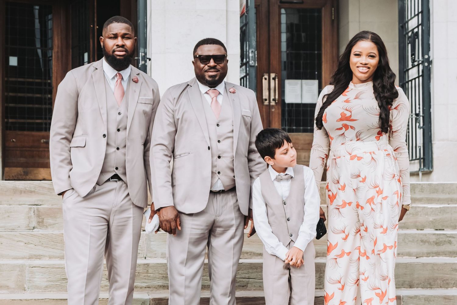 Bridal party in light gray suits and floral print dresses stand on stone steps for photos.