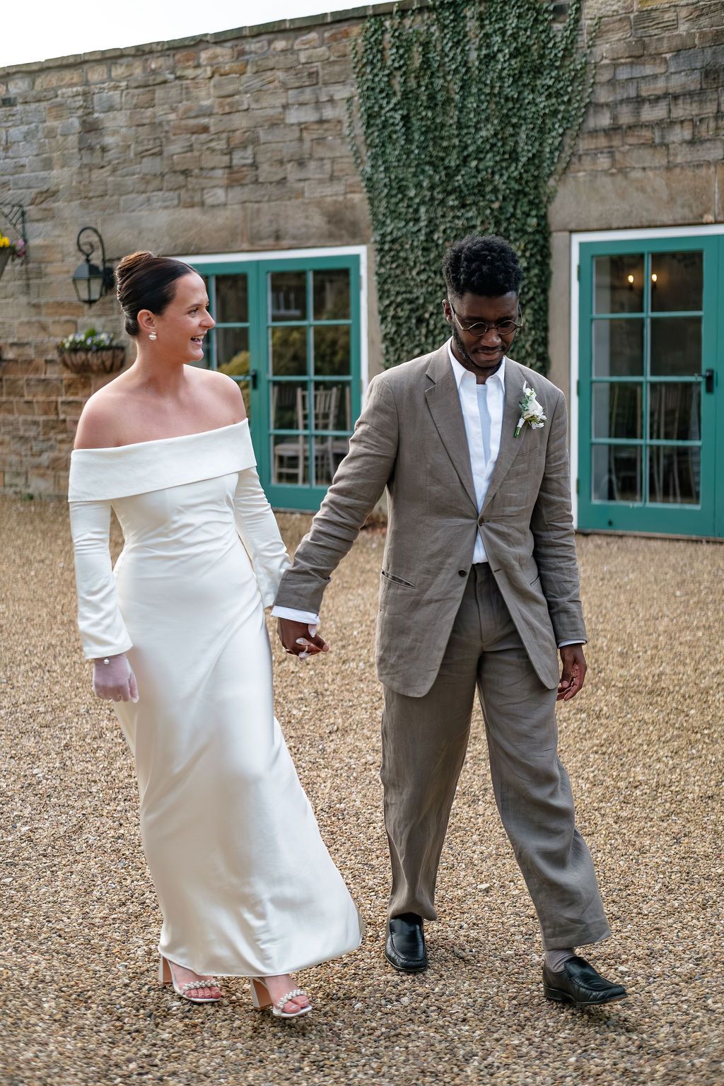 A bride in white off-shoulder gown and groom in tan suit hold hands walking outside a stone building.