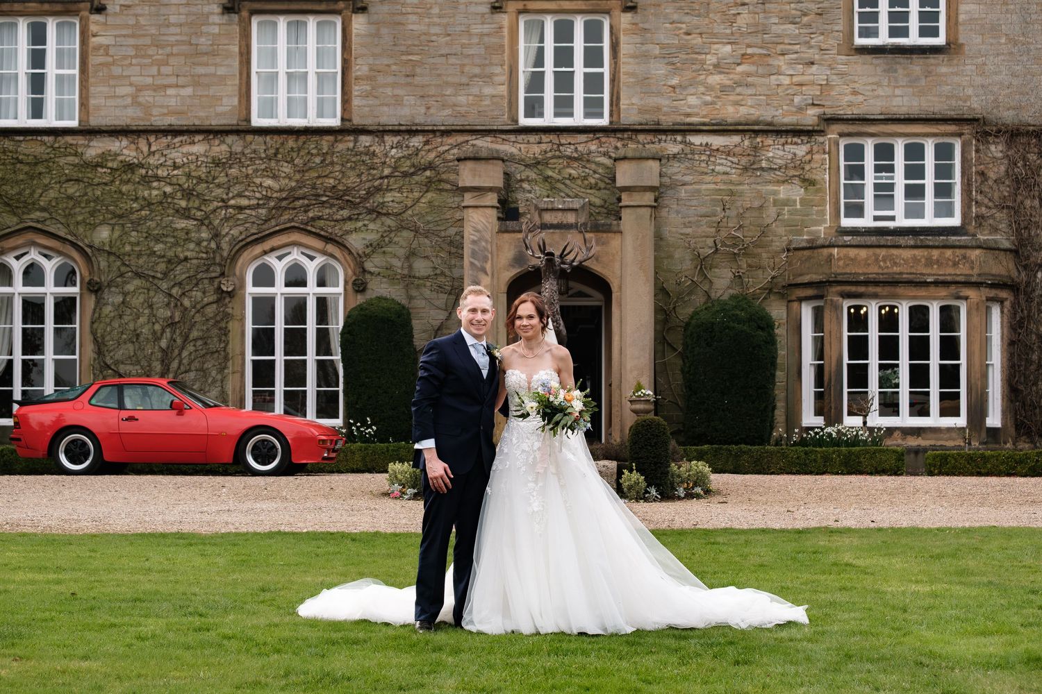 Bride and groom posing outside a stone manor house with a red sports car, bride in a tulle gown holding a bouquet.