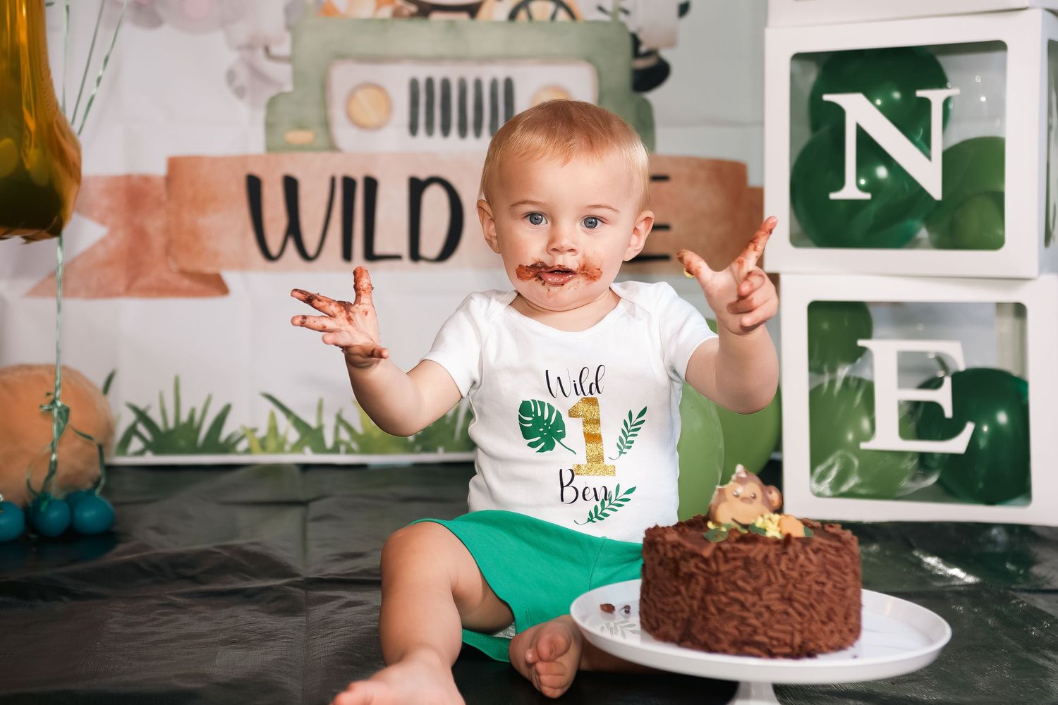 A young child wearing a 'Wild One' shirt enjoys a chocolate cake during their first birthday celebration.