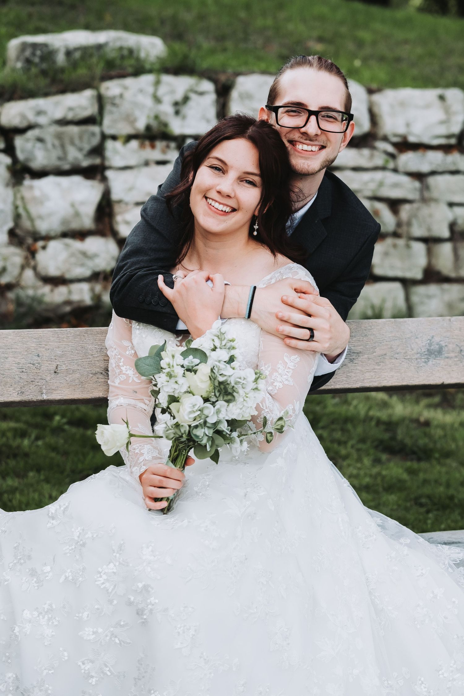 A romantic outdoor wedding portrait features a white bouquet and lace dress against a stone wall.