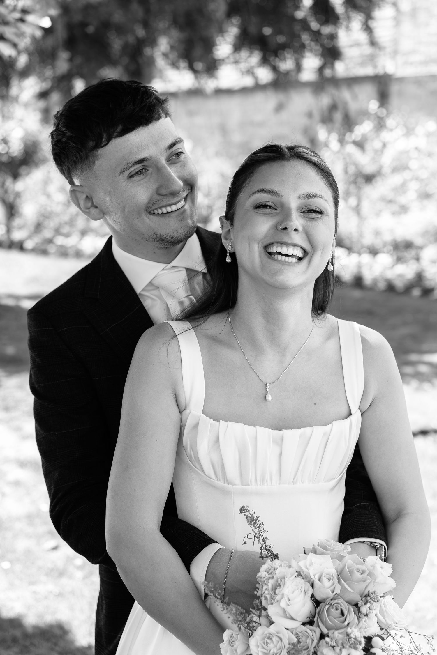 Black and white photo of a smiling bride holding flowers and groom embracing outdoors on their wedding day.