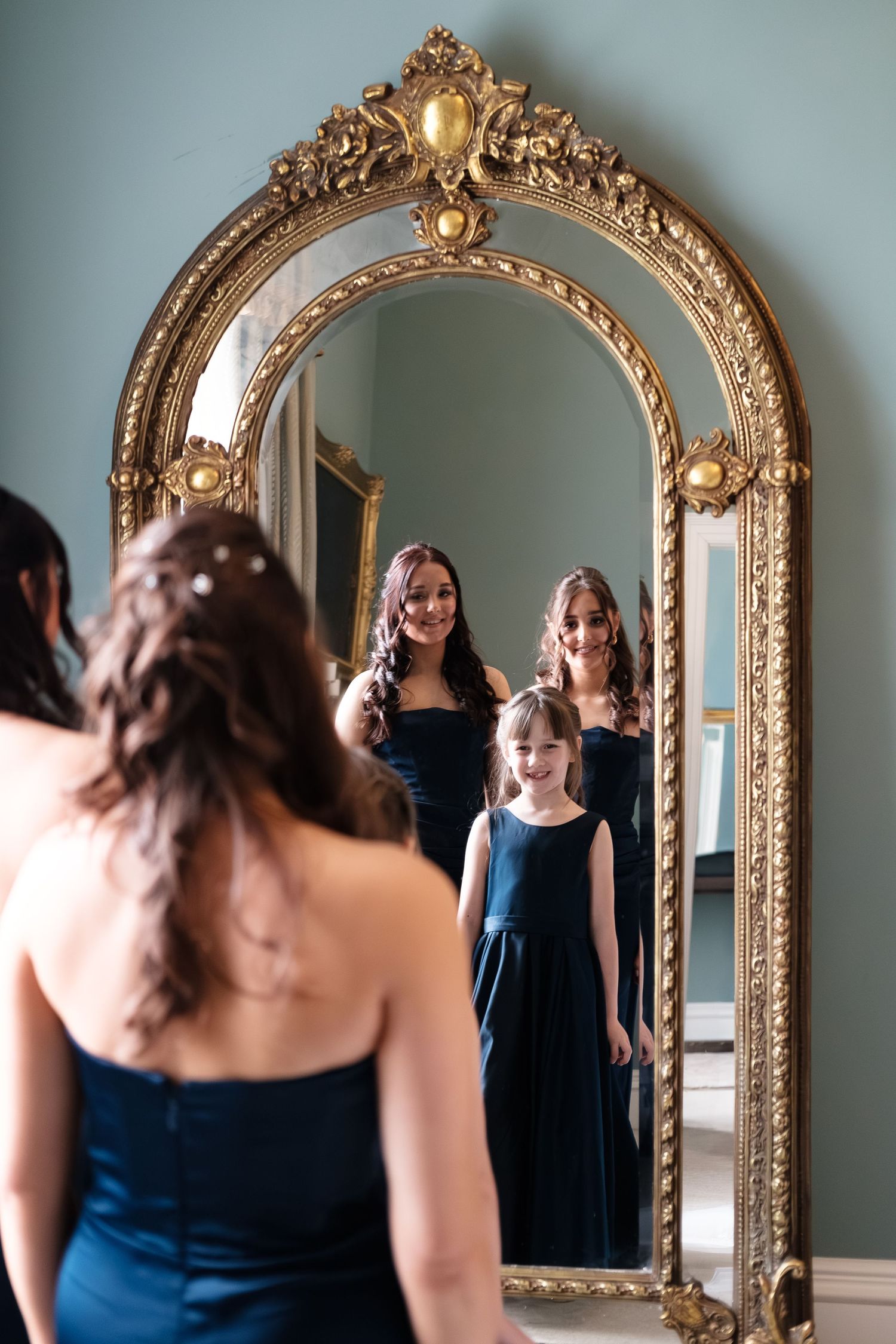 Bridesmaids in navy blue dresses reflected in an ornate gold mirror during wedding preparations.