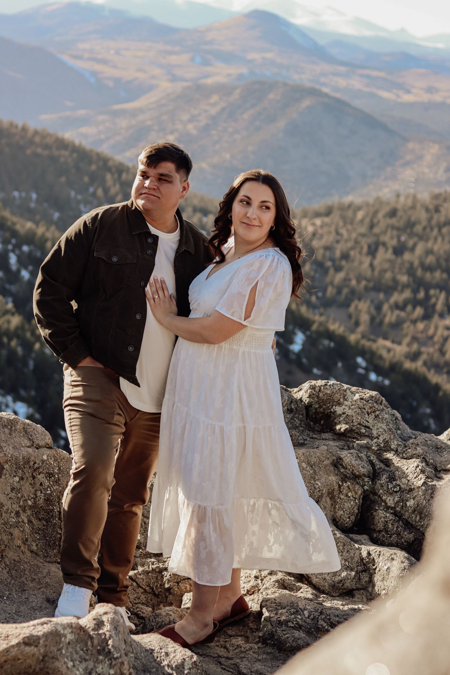 Couple posing on rocky mountain overlook, woman in white dress, man in dark jacket, scenic Colorado mountains backdrop.