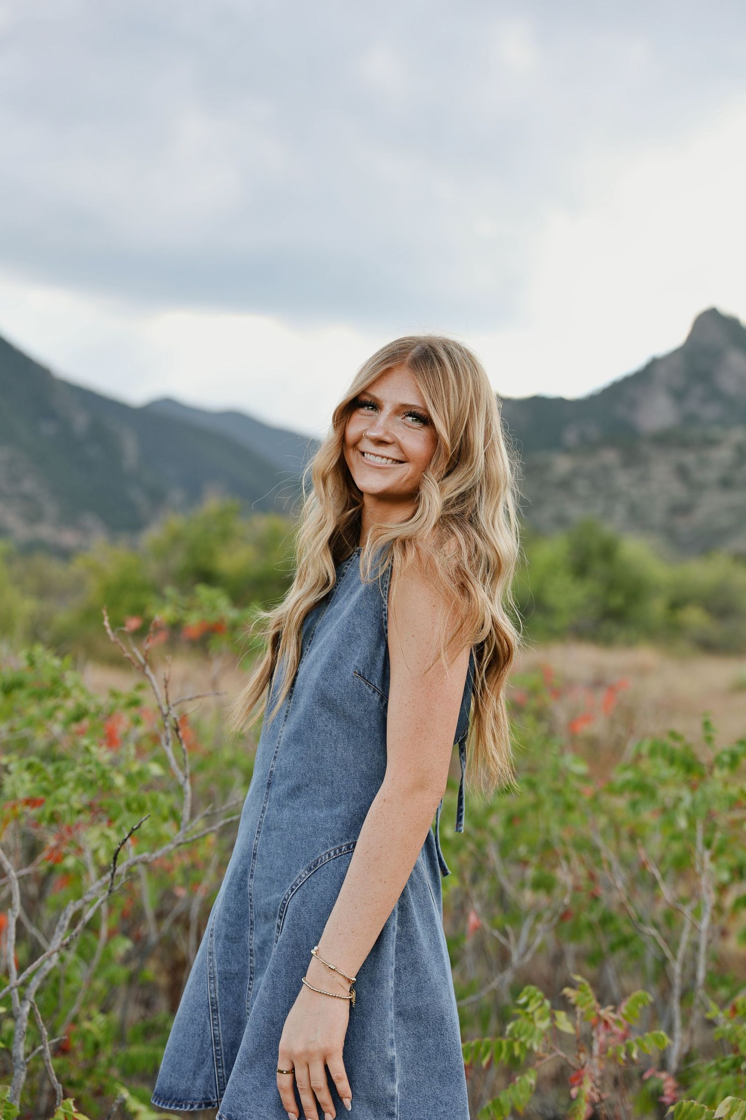 Smiling blonde woman in denim dress poses in mountain meadow with green shrubs and cloudy sky backdrop.