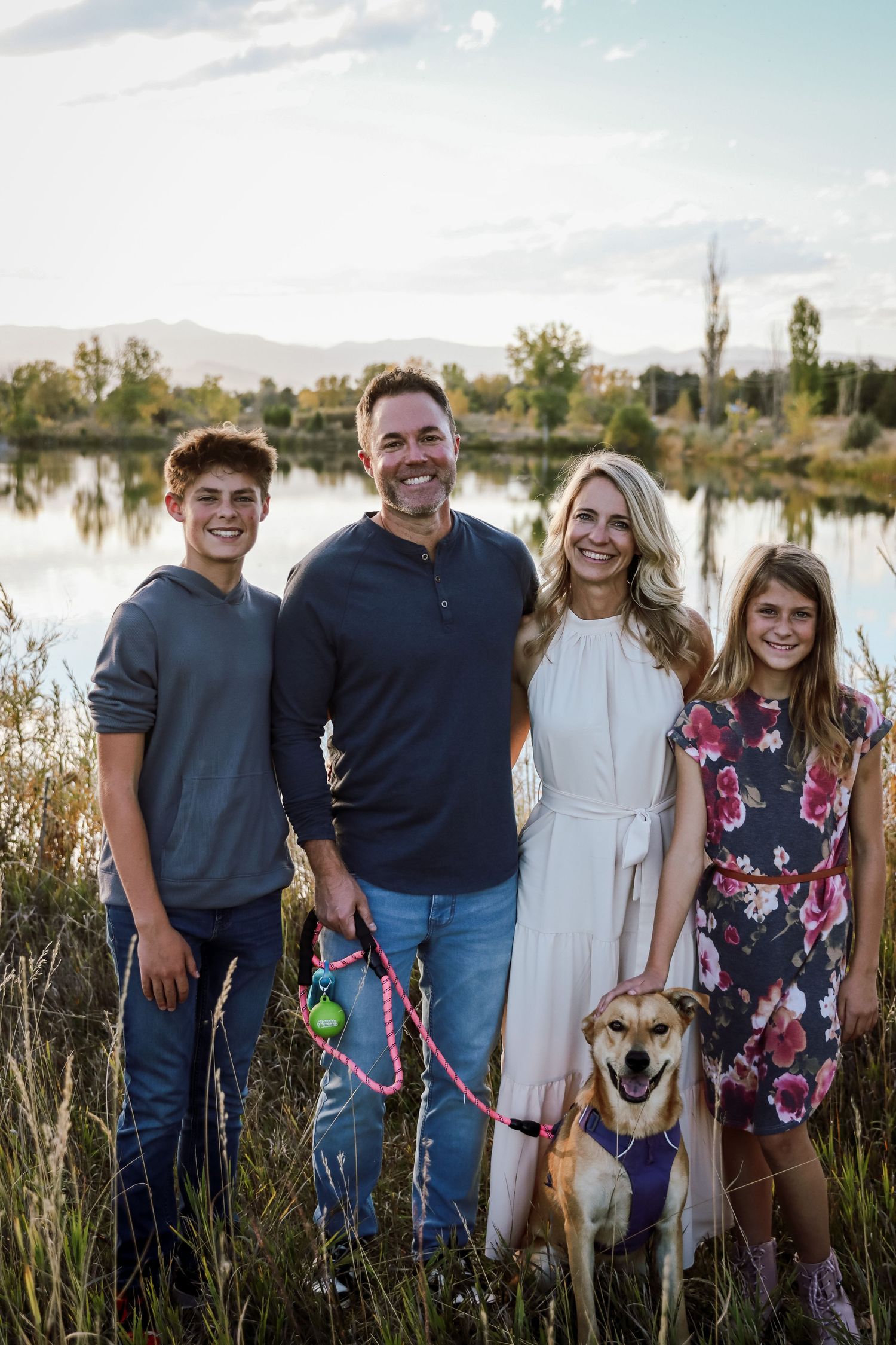 Happy family of four posing outdoors with their golden dog near a scenic lake and mountains at sunset.