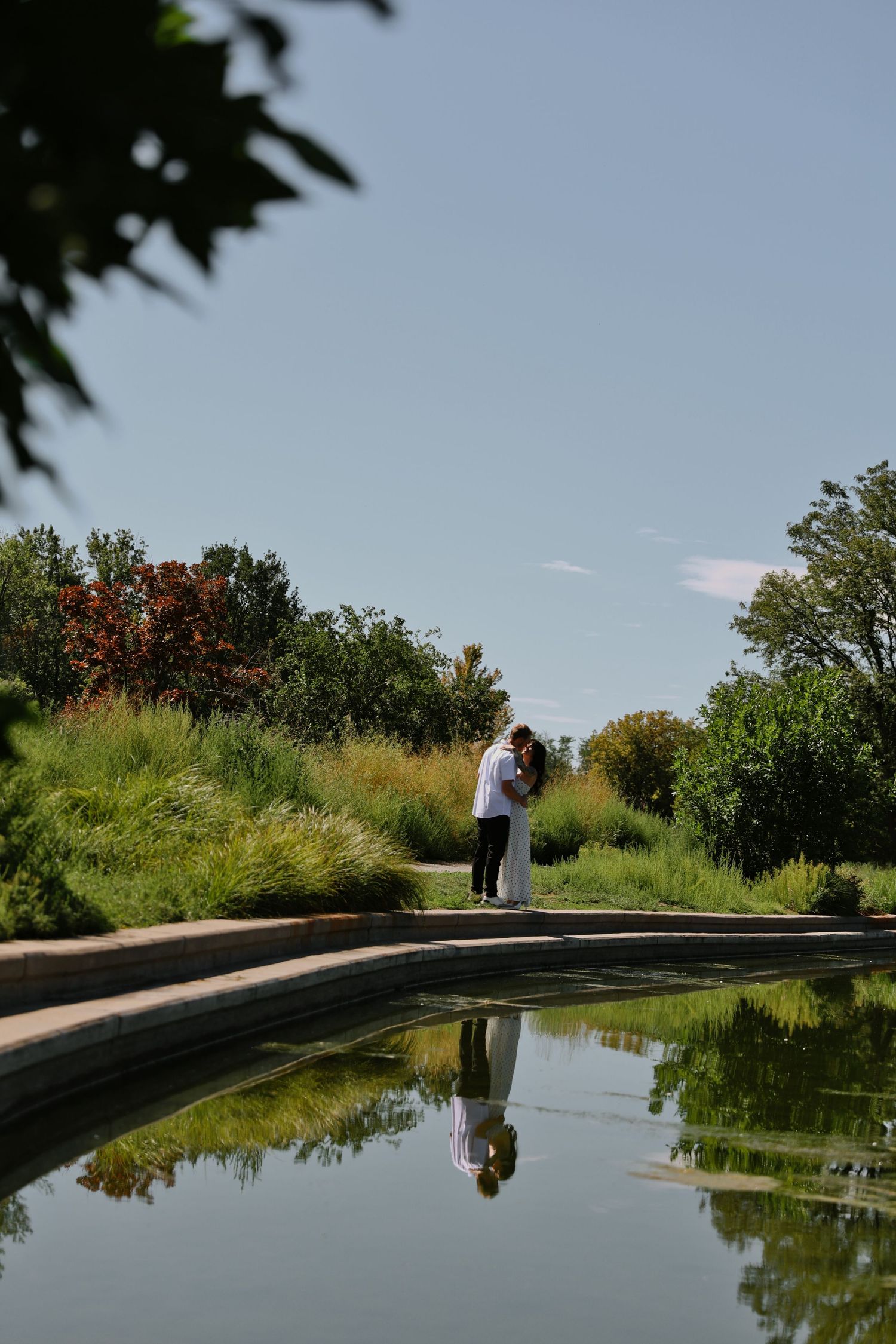 Couple sharing a romantic kiss beside a reflective garden pond, surrounded by lush greenery under a clear blue sky.