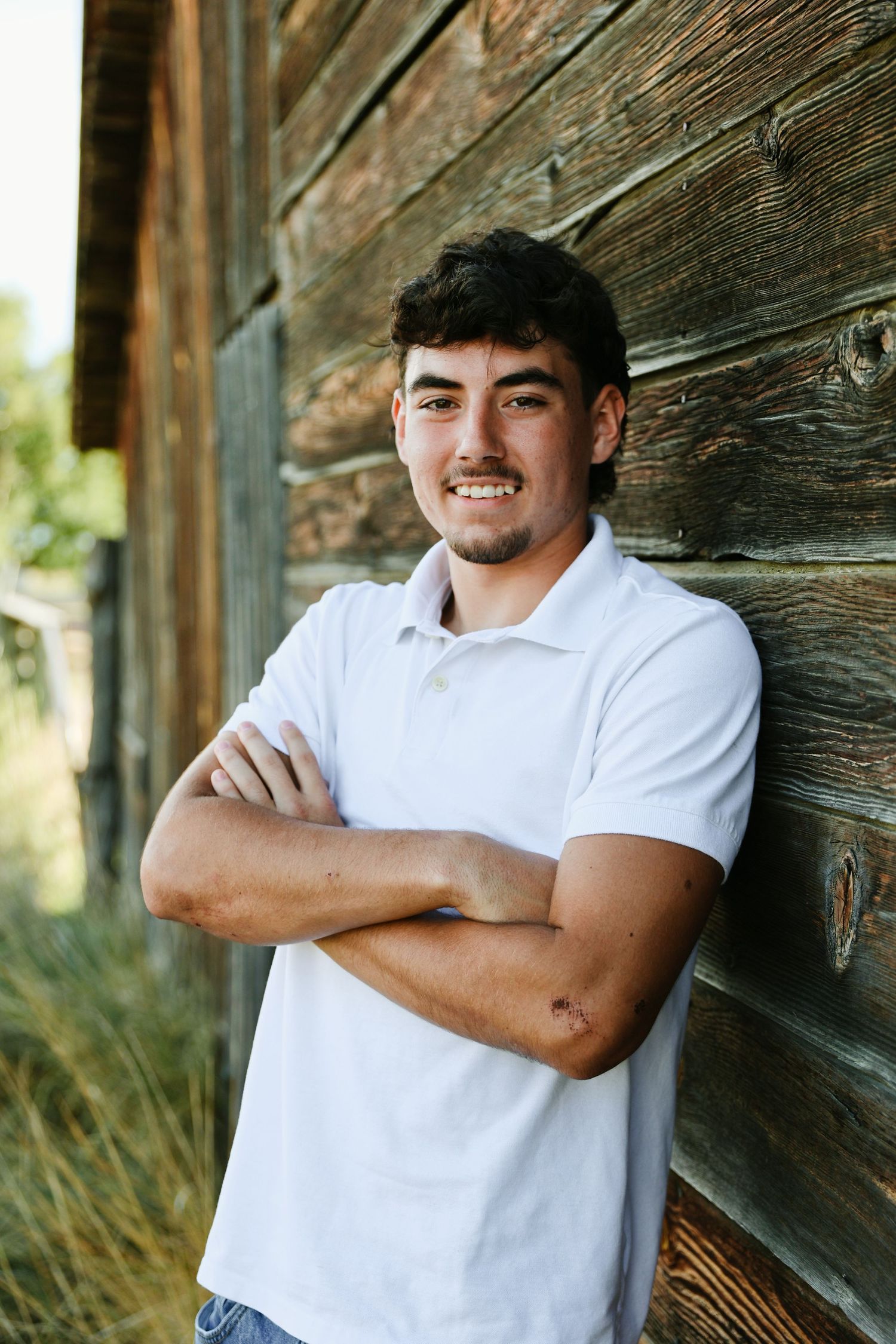 Young man in white polo shirt smiling with arms crossed, leaning against rustic wooden wall outdoors.