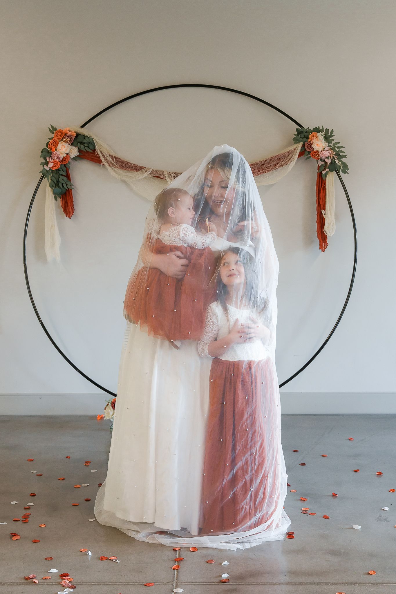 Two figures share an intimate moment under a sheer veil with floral decorations and scattered rose petals.