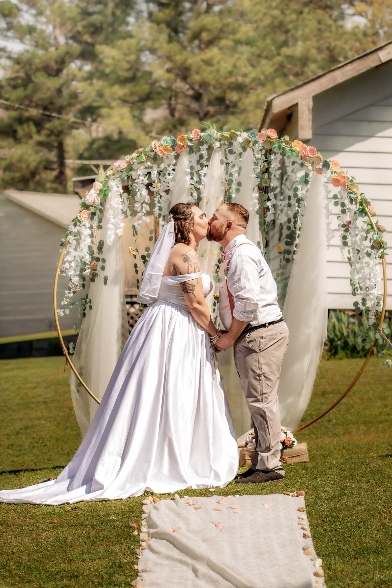 Bride and groom share first kiss under floral hoop arch at outdoor backyard wedding ceremony.