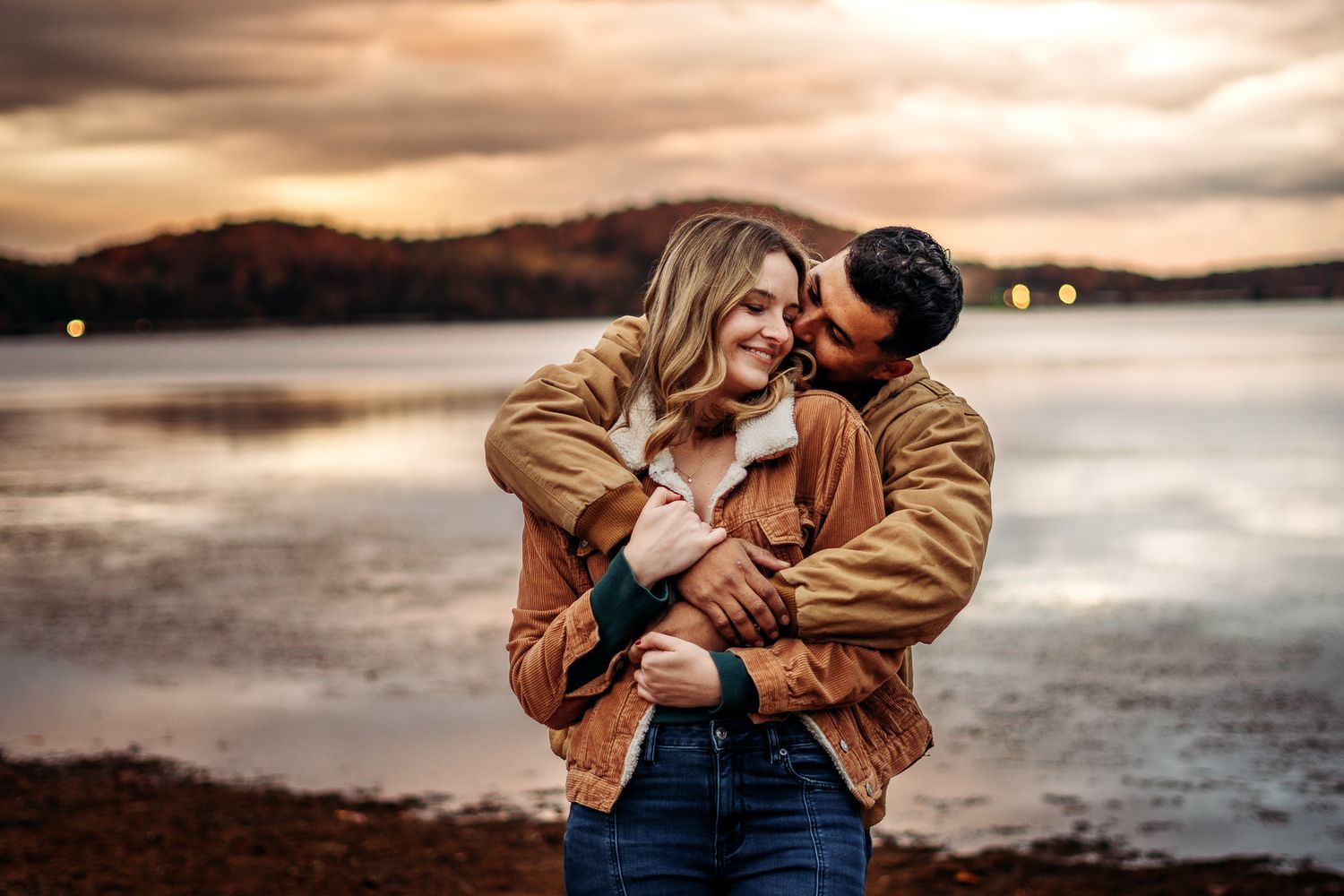 Romantic couple embraces on a beach at sunset with dramatic cloudy sky and water in the background.