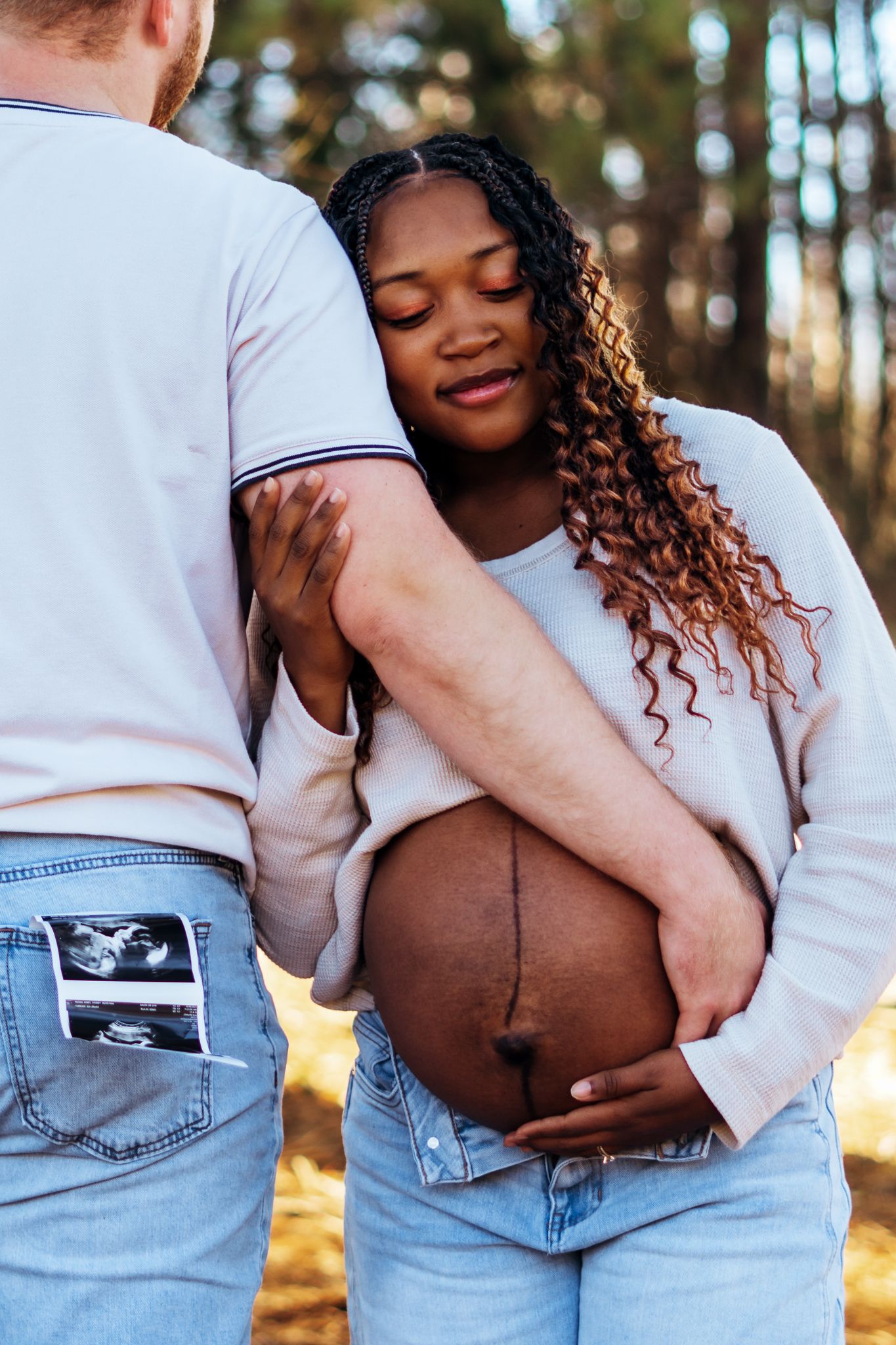 Couple holding ultrasound photo in their back pocket while embracing outdoors in a sunlit forest setting.