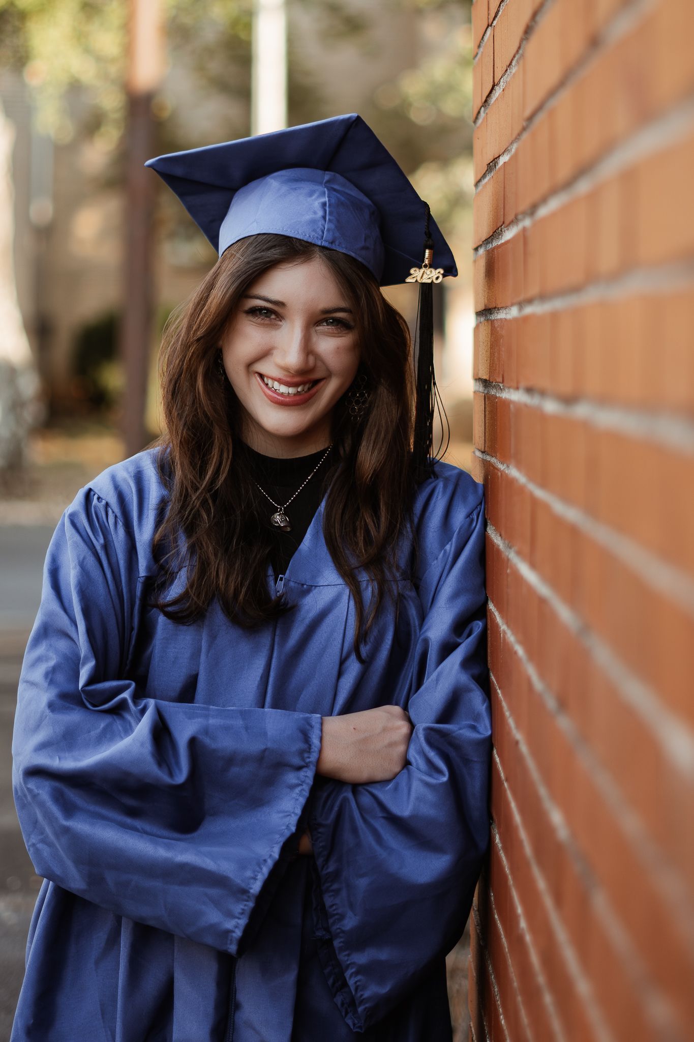 Graduate in blue cap and gown poses smiling against brick wall.