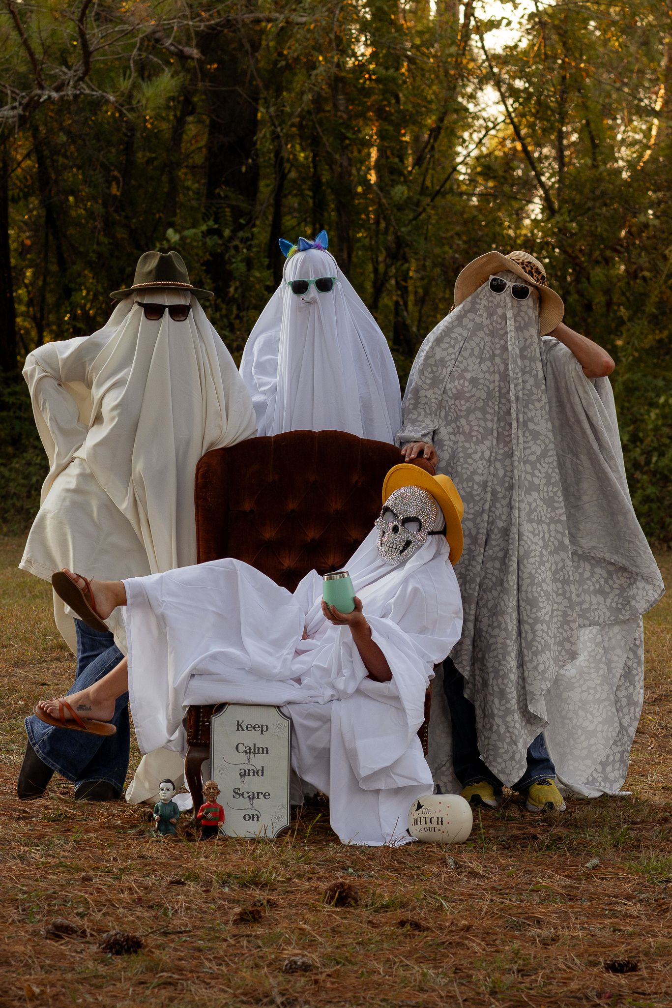 People dressed as ghosts with white sheets pose with a vintage brown sofa in a wooded setting.