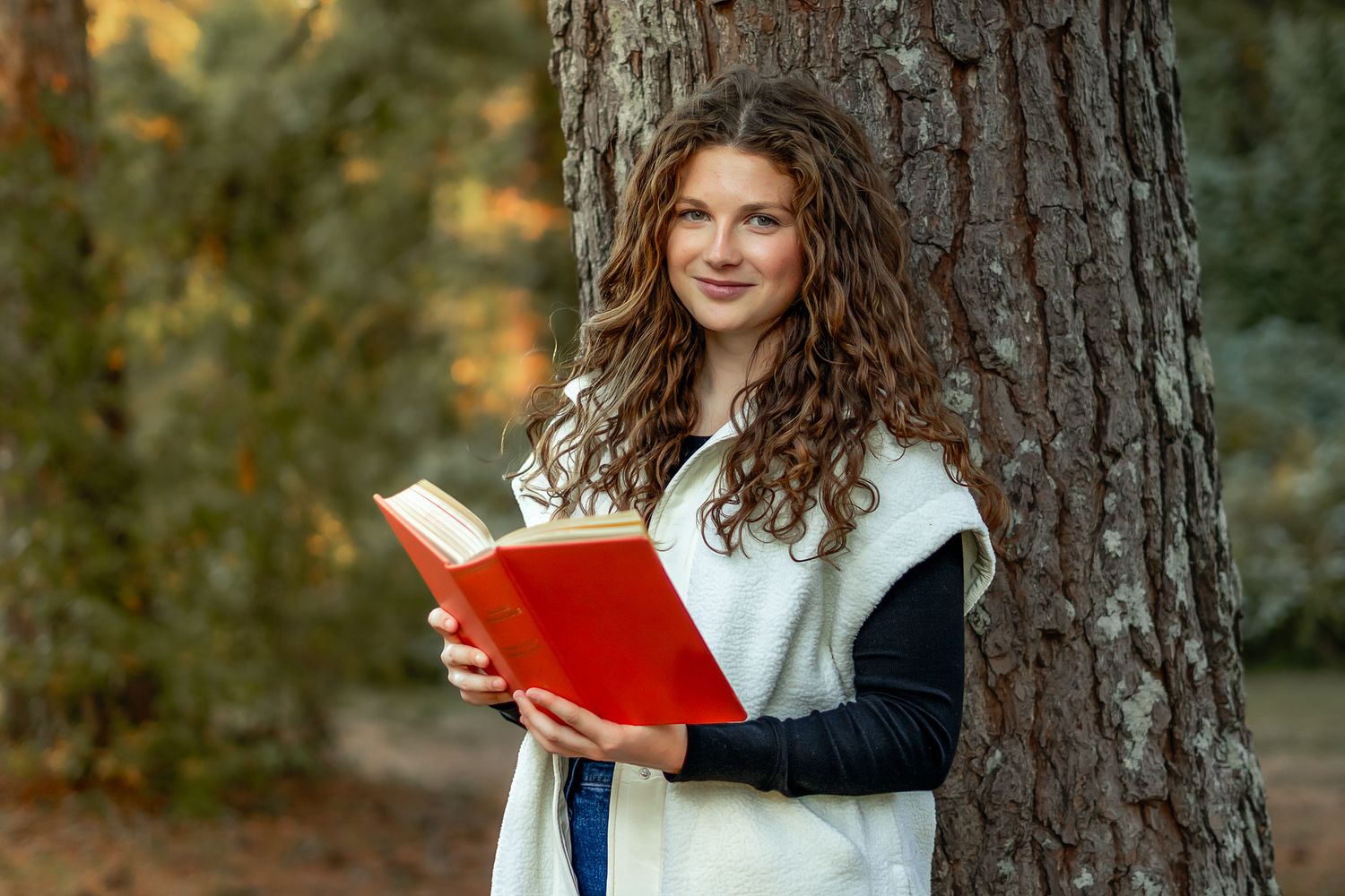 Student reading red book while standing by large tree trunk in autumn setting.