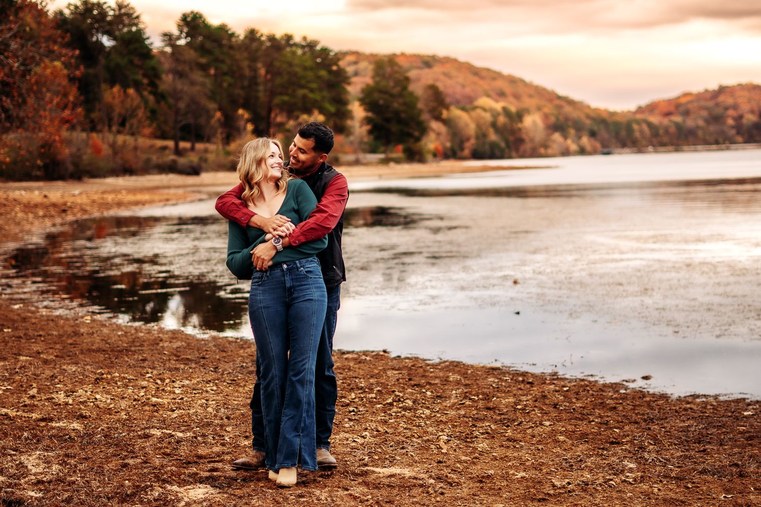 A couple shares a romantic embrace at sunset by a serene lake with autumn foliage in the background.