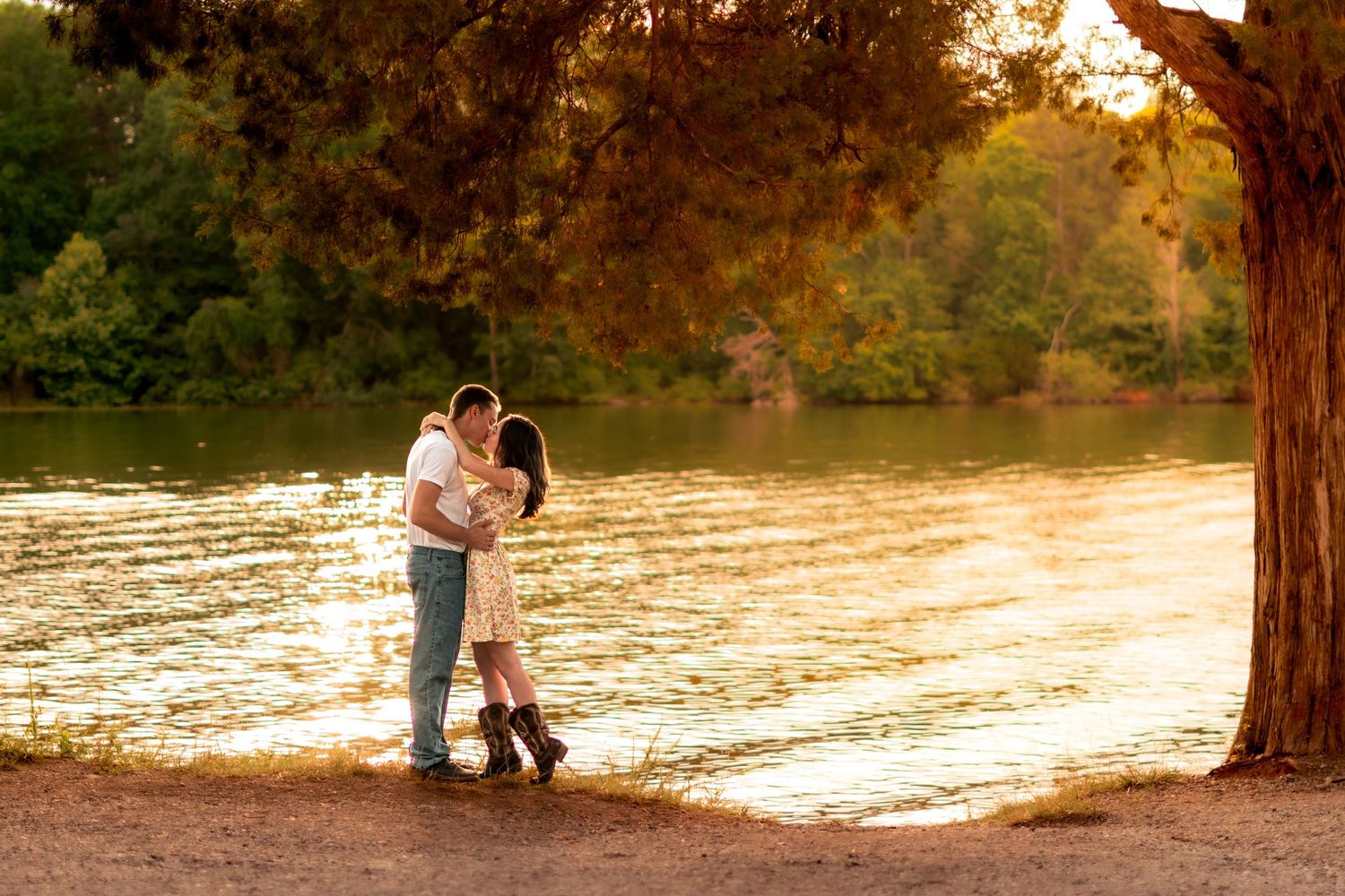 Couple sharing a romantic kiss by a sunlit lake under a large tree during golden hour in a park.