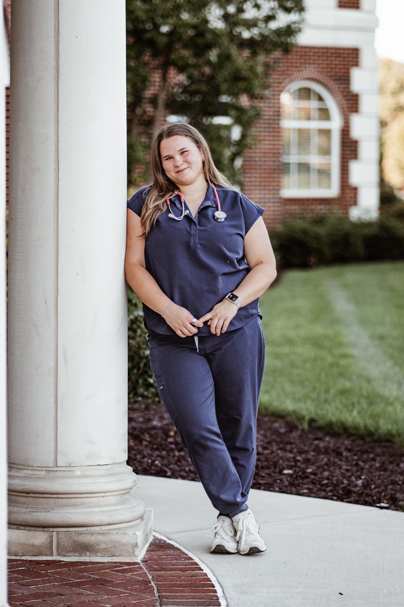 Nursing student in scrubs poses confidently by white column on campus.