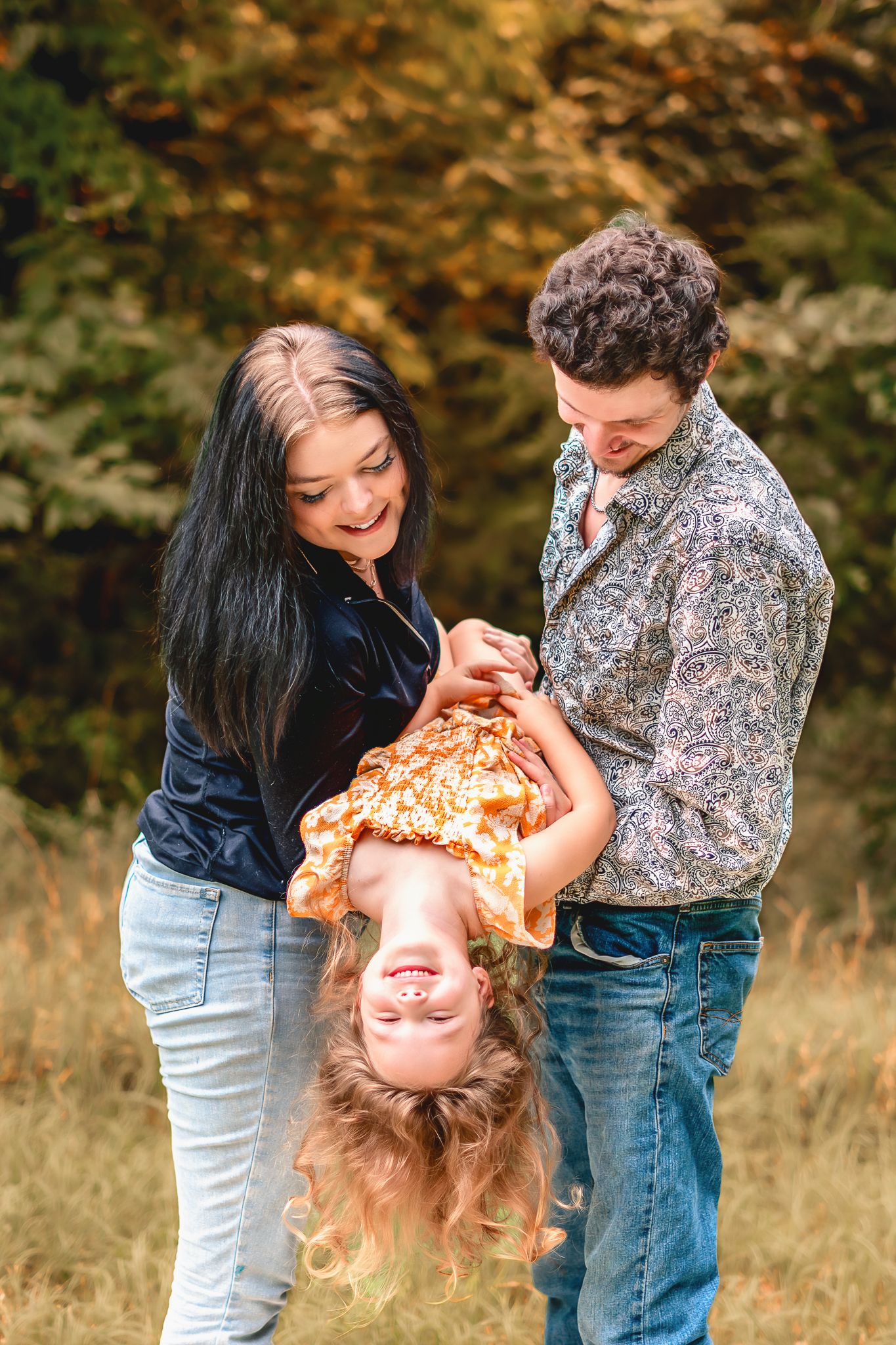 A joyful family plays outdoors in autumn, parents swinging their laughing child upside down in a golden fall setting.