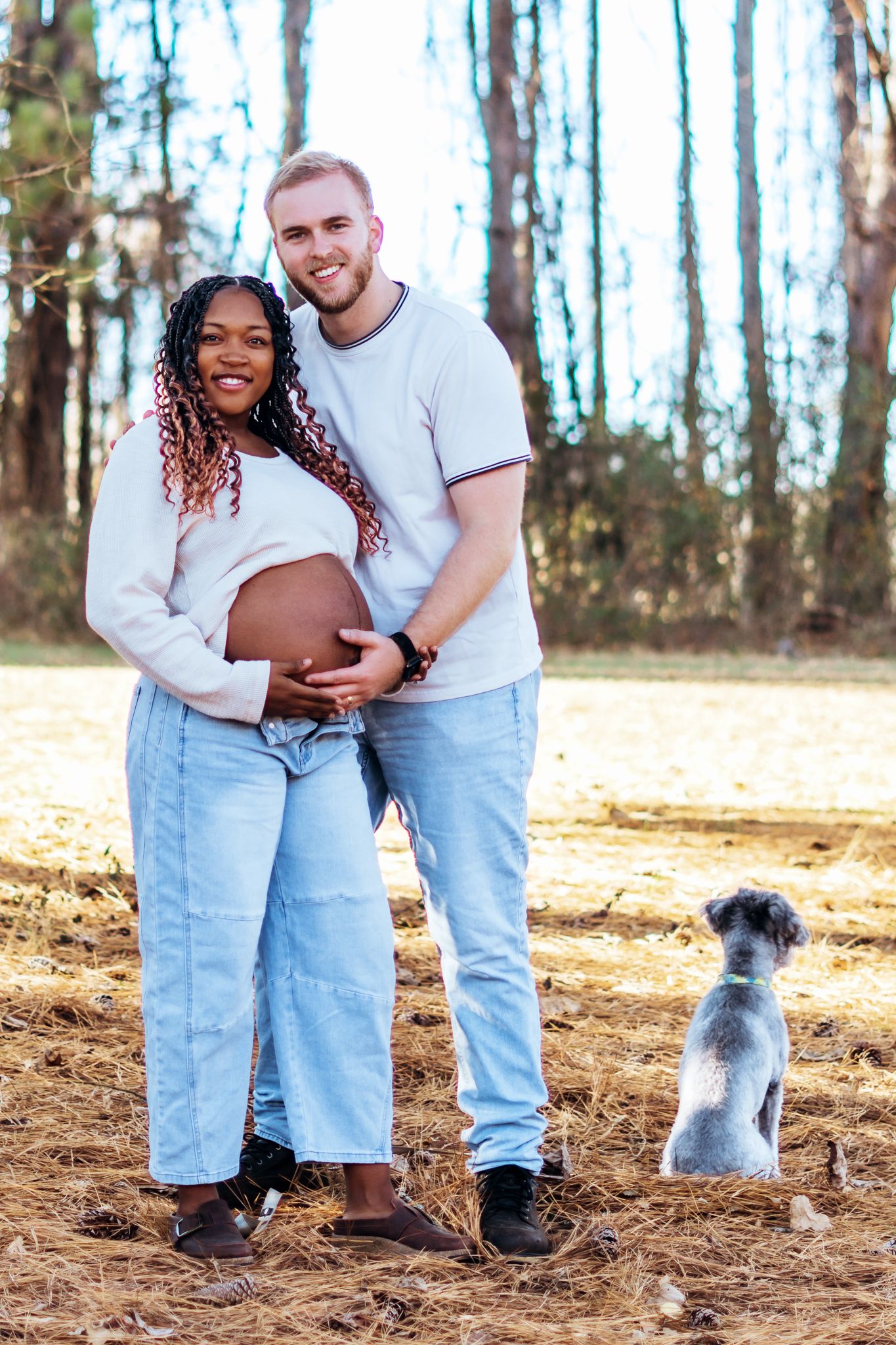 A couple in casual outfits stands together in a wooded area with their small dog, holding a basketball.