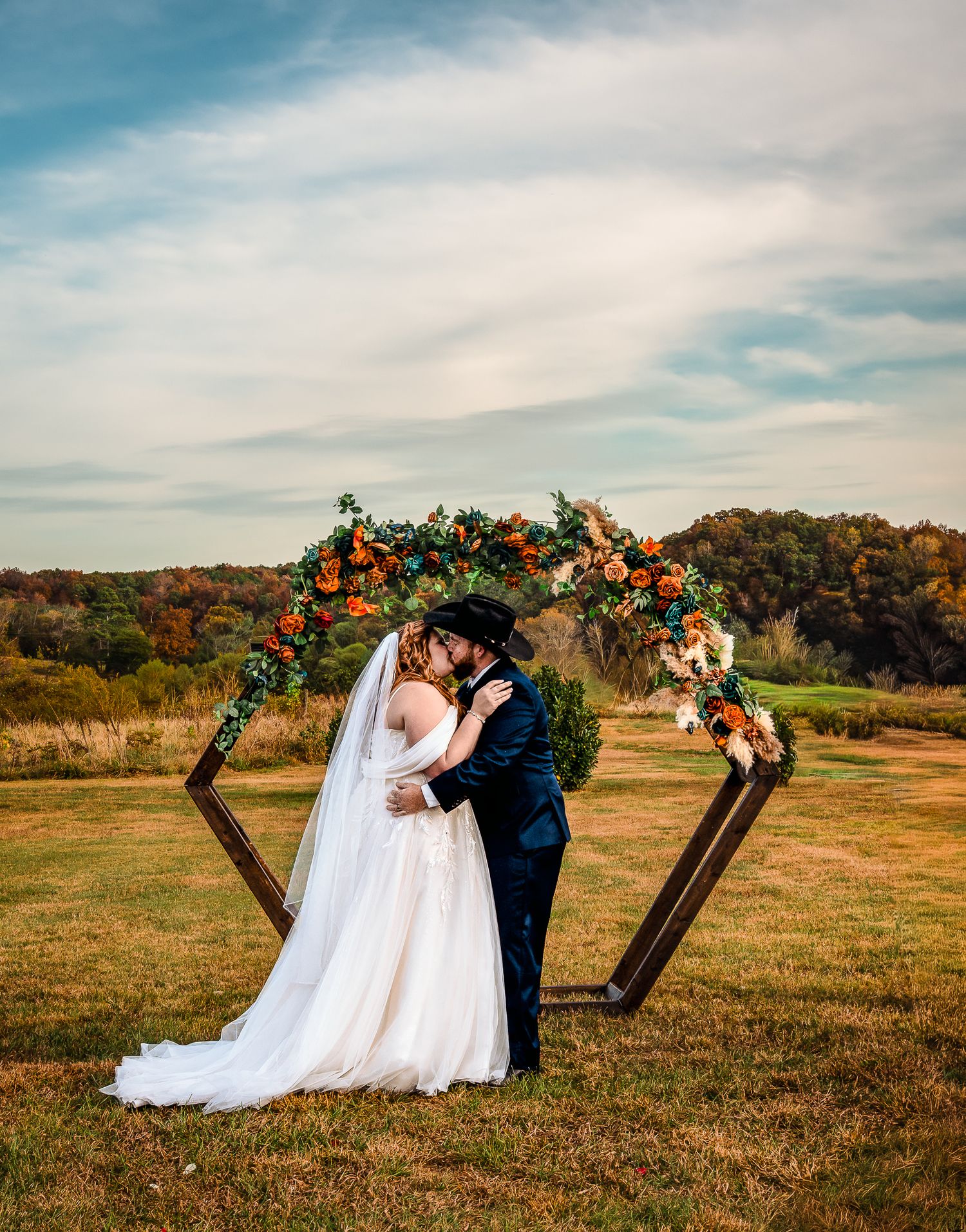 Bride and groom share first kiss under a geometric wood arch adorned with orange and green florals at an outdoor fall wedding.