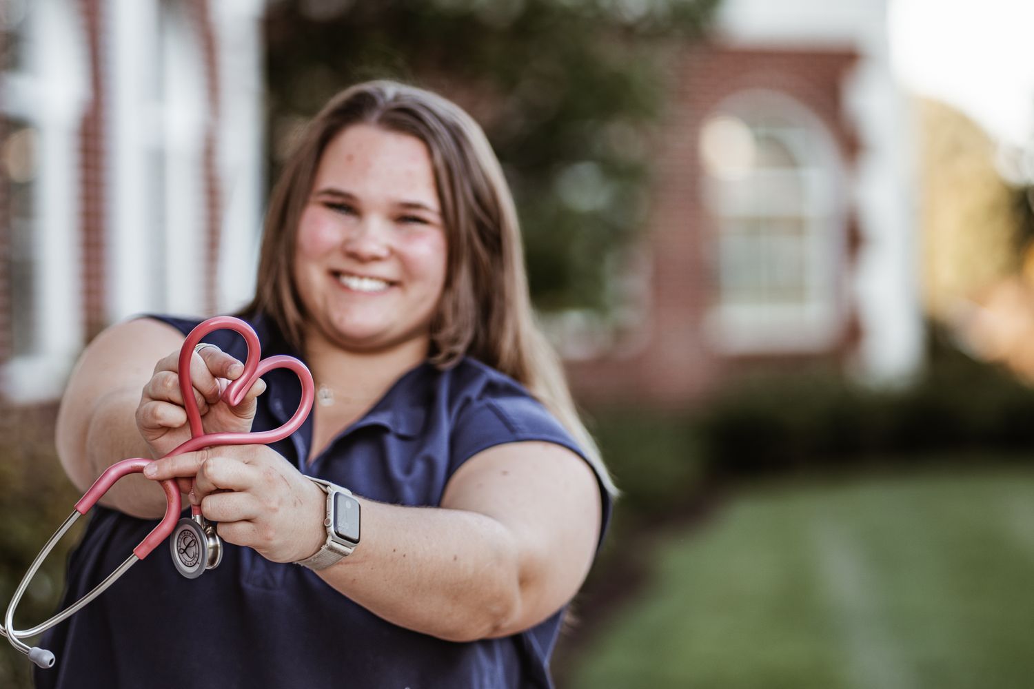 Student holding a pink stethoscope while wearing a navy shirt outdoors.