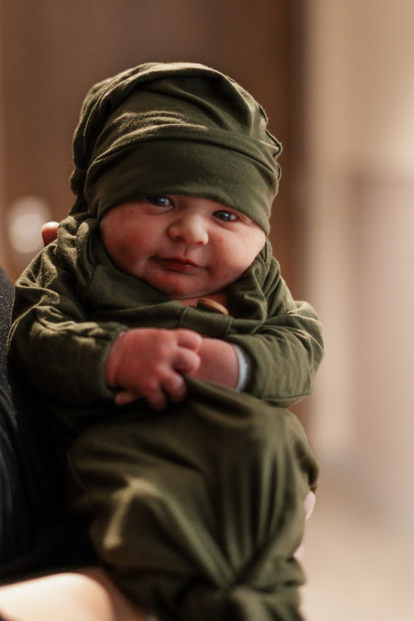 A small baby wrapped in olive green fabric with eyes peeking out.