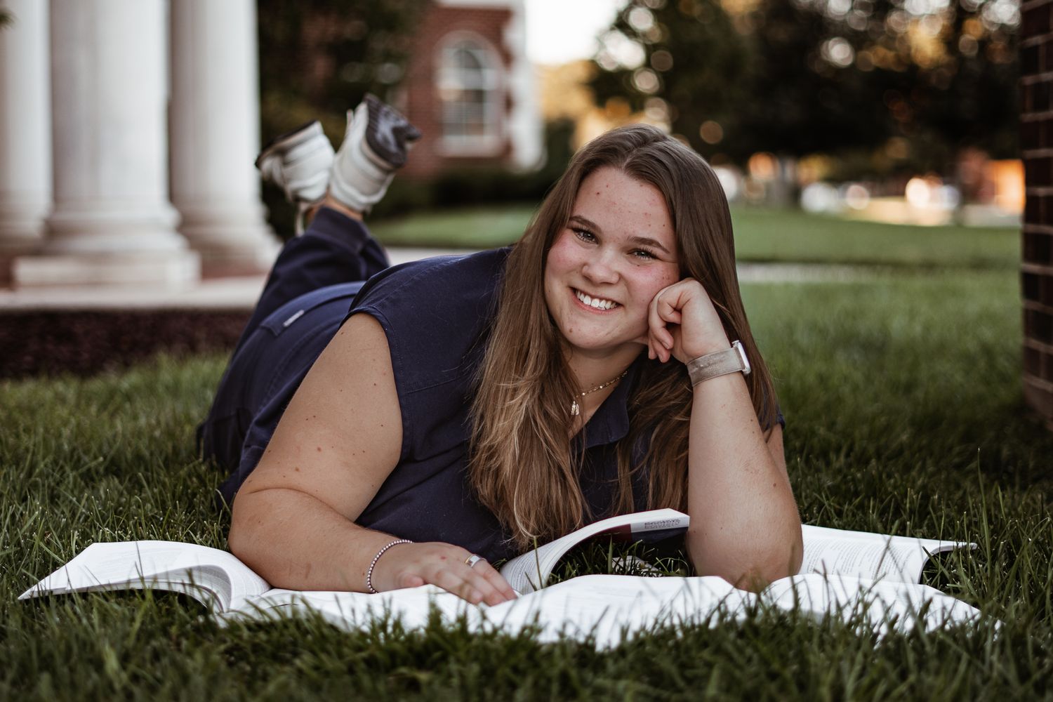 Student lying on grass reading a book outdoors near white columns during golden hour.