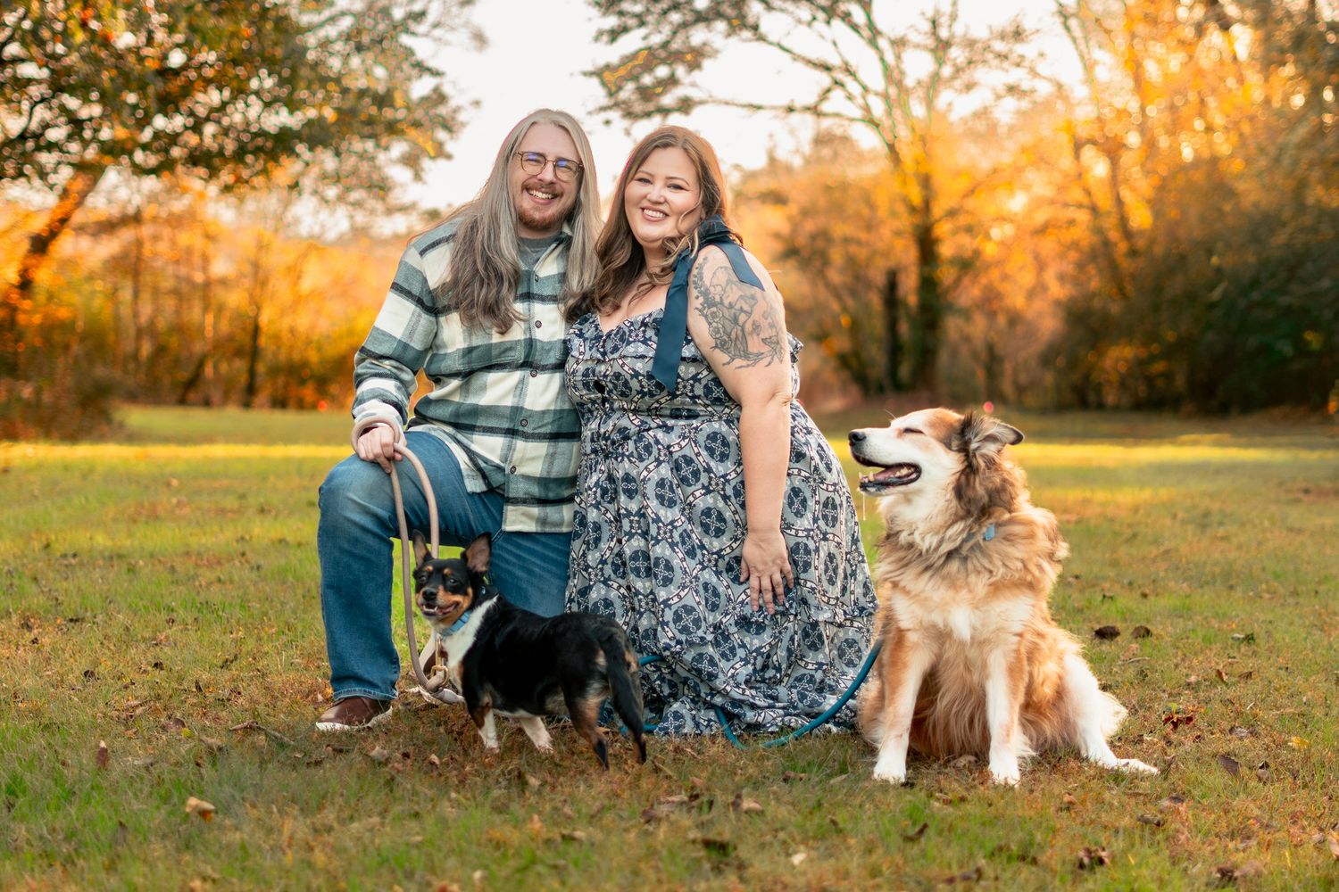 Two people and a dog sit together in a grassy field at sunset during an autumn photoshoot.