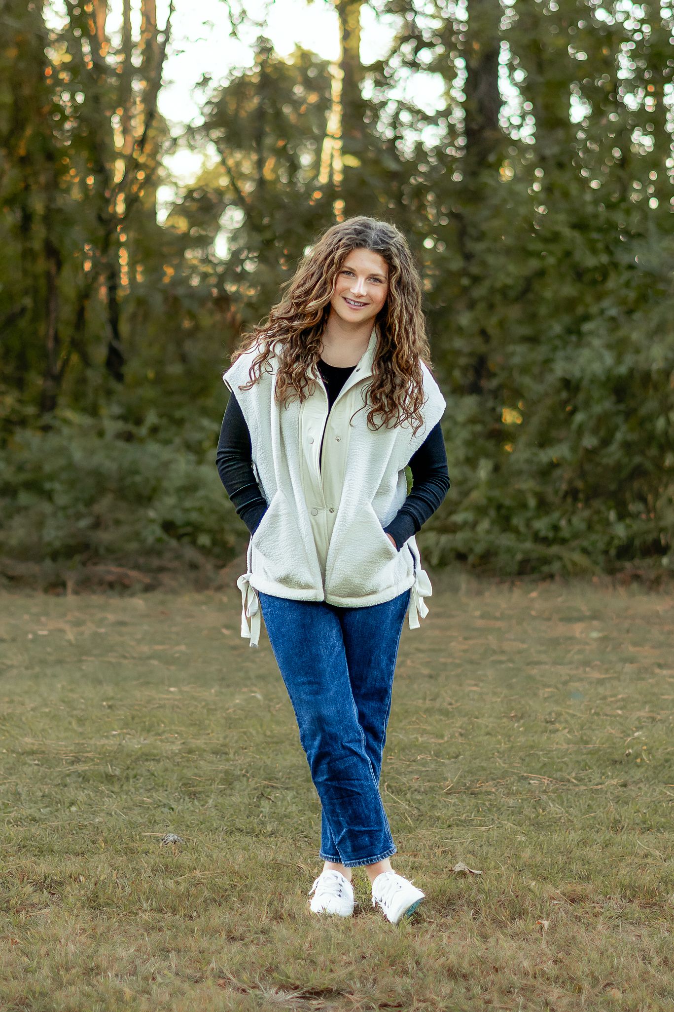 Student wearing white and navy jacket with jeans standing in wooded area.
