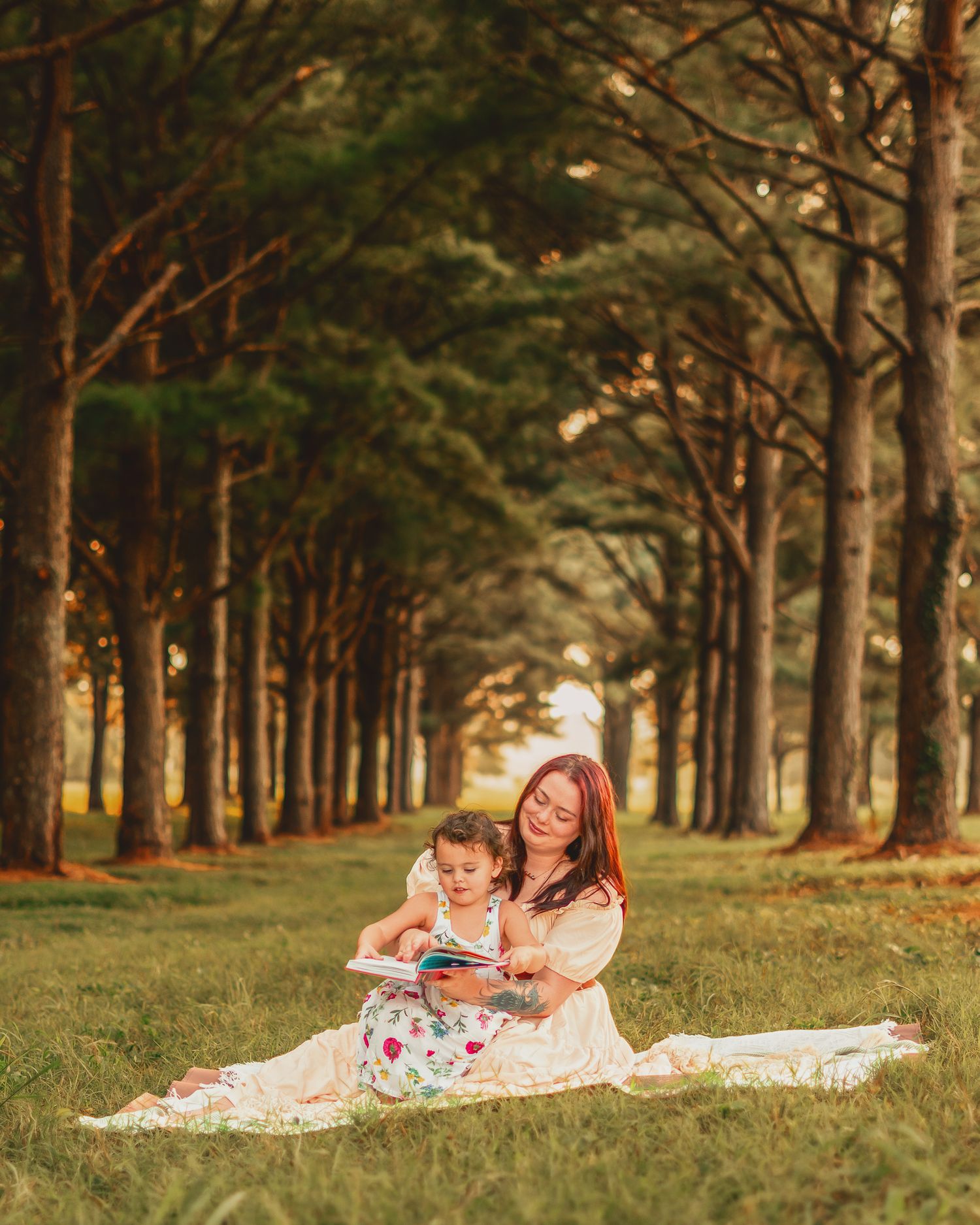 A mother and toddler sit on a blanket reading a book together in a lush green tree-lined park.