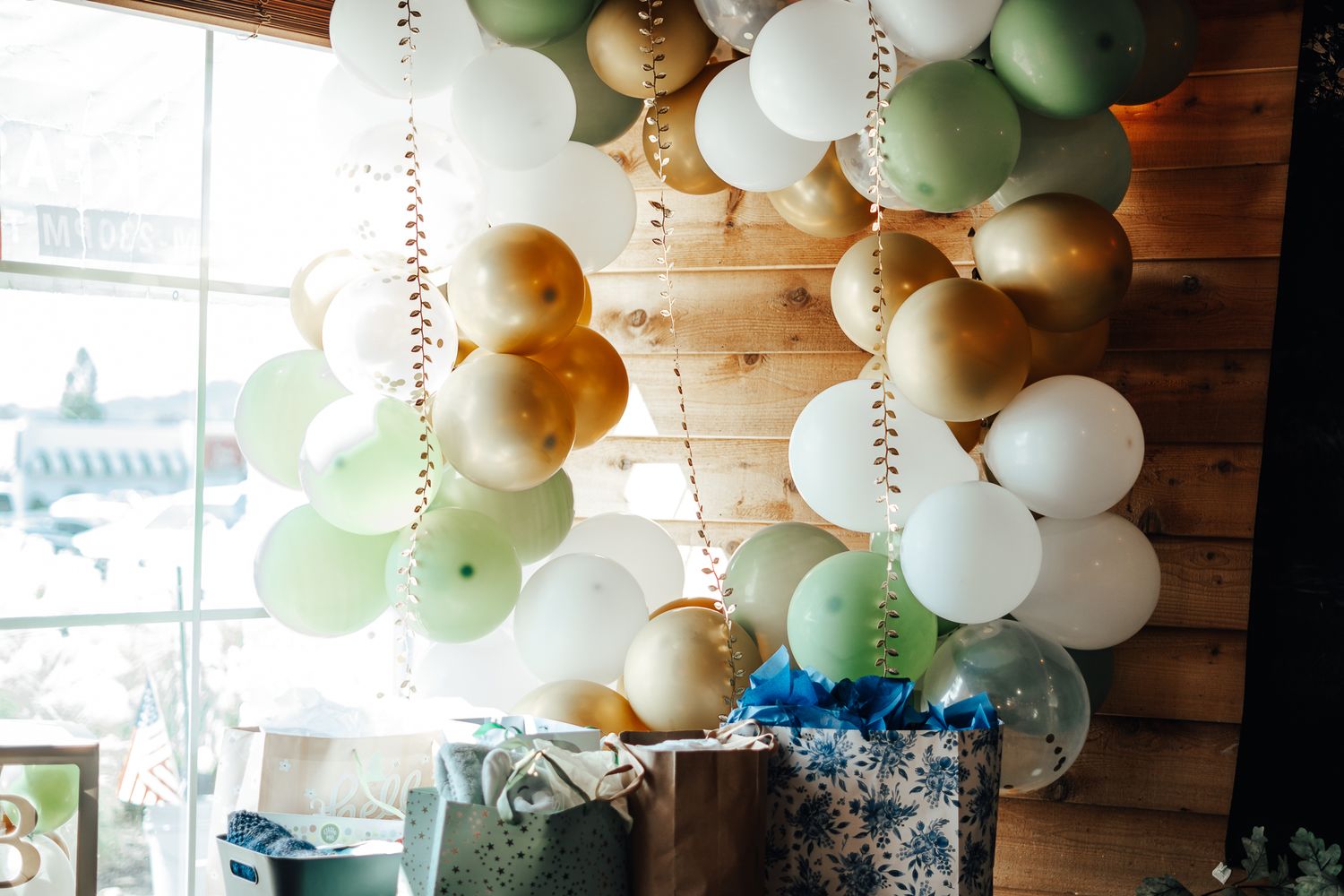 Elegant balloon garland with sage green, gold, and white balloons decorates a rustic wooden wall.