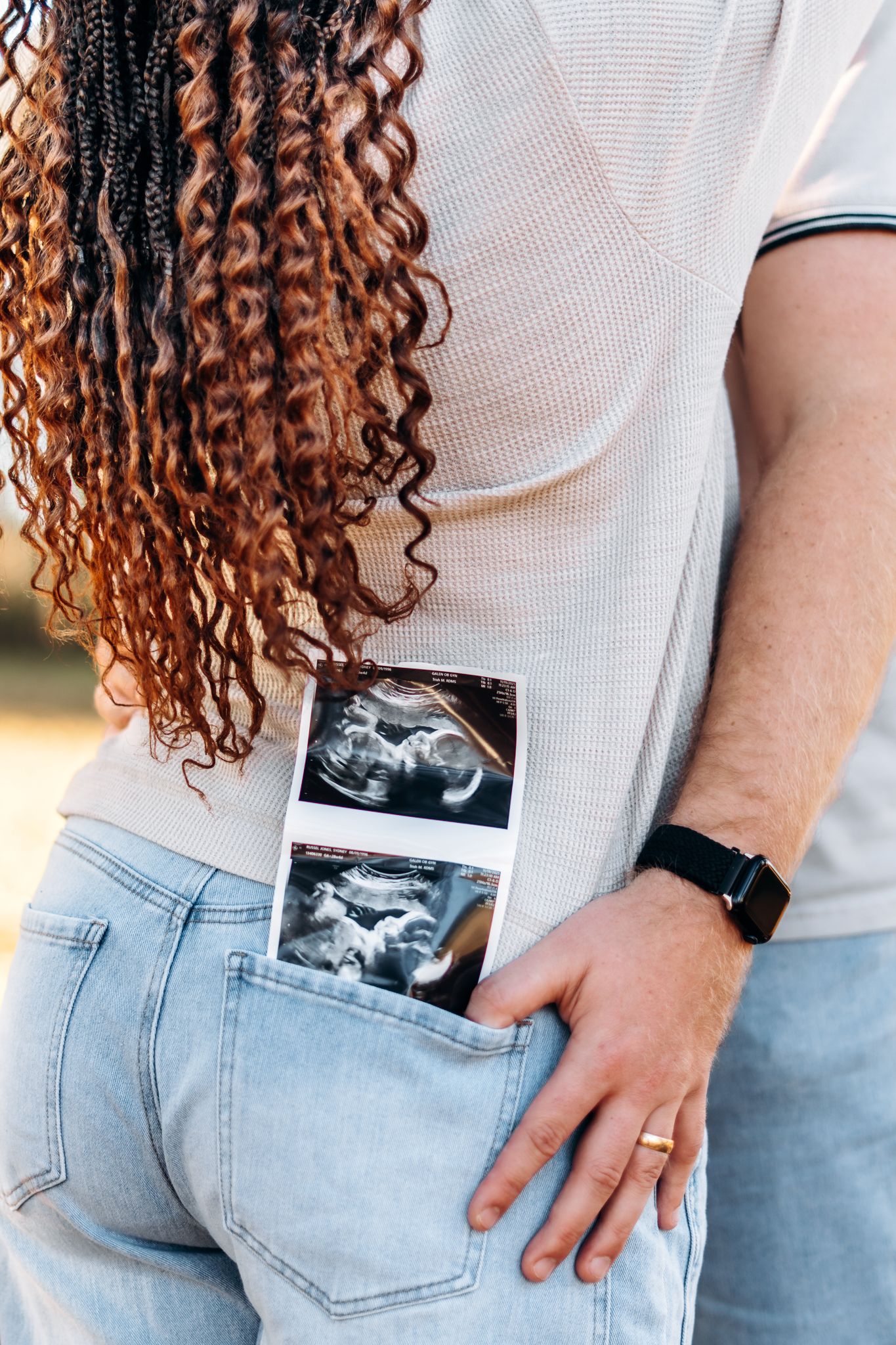 Ultrasound photos tucked into back pocket of jeans with curly copper-toned hair visible in white shirt.