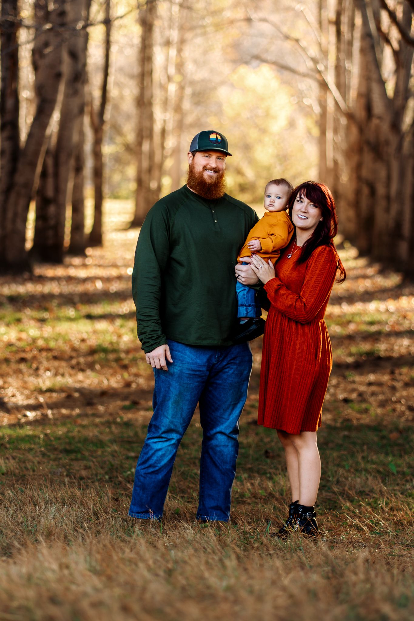 A couple stands together in a wooded area during autumn, with fallen leaves scattered on the ground.