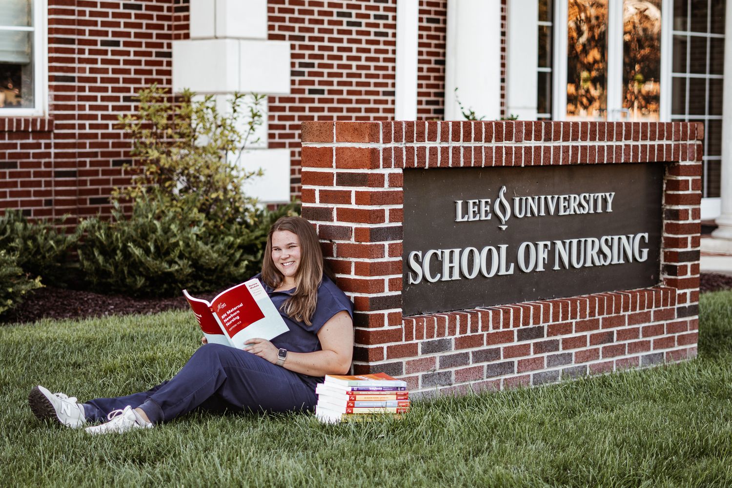 Student reading book while sitting by Lee University School of Nursing brick sign.