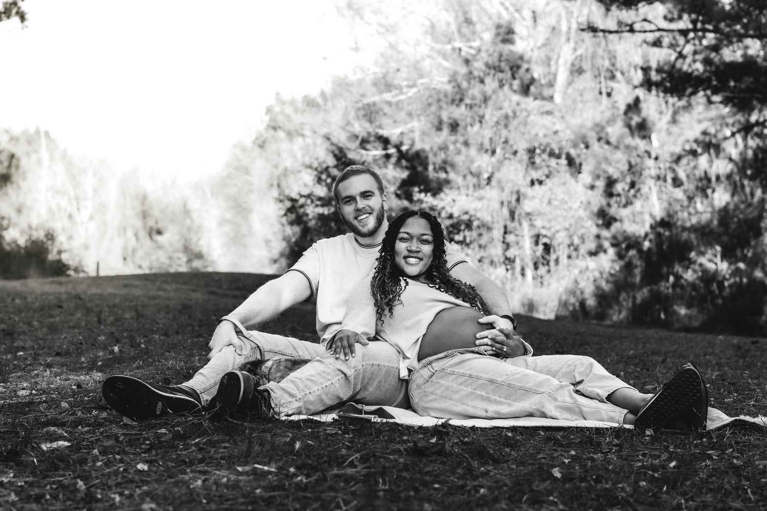 Black and white photo of couple relaxing together on blanket in grassy field with pine trees.