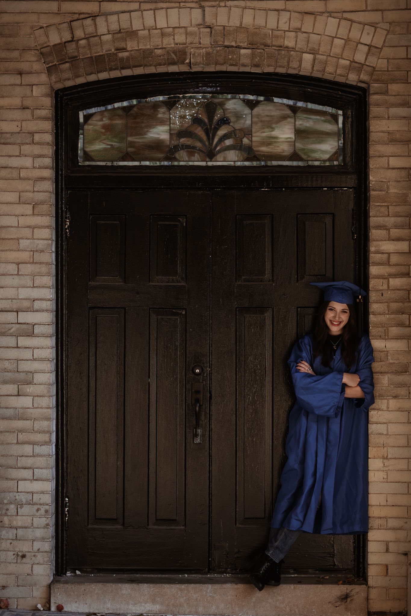 Graduate in blue regalia poses artistically by dark wooden door with brick archway.