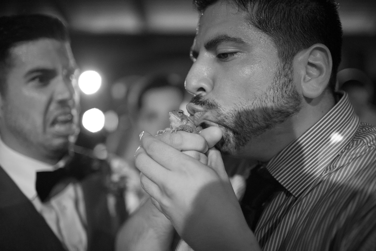 Black and white photo of a man eating food at a formal event with a bow-tied guest in the background.