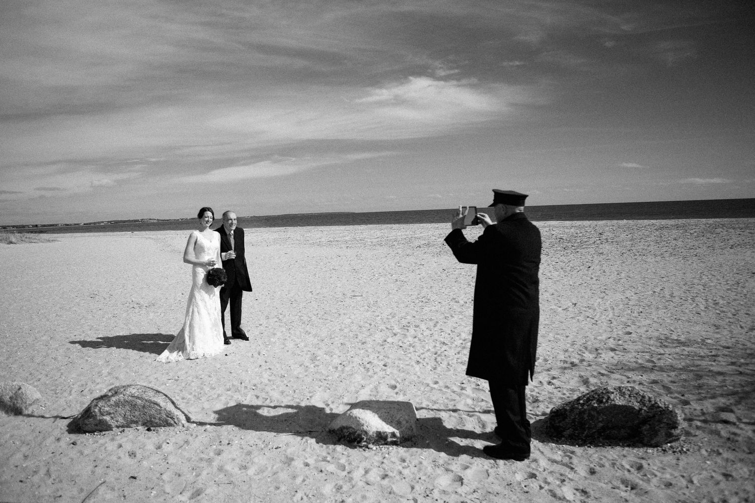 Black and white beach wedding ceremony with bride, groom, and officiant on sandy shore under cloudy sky.